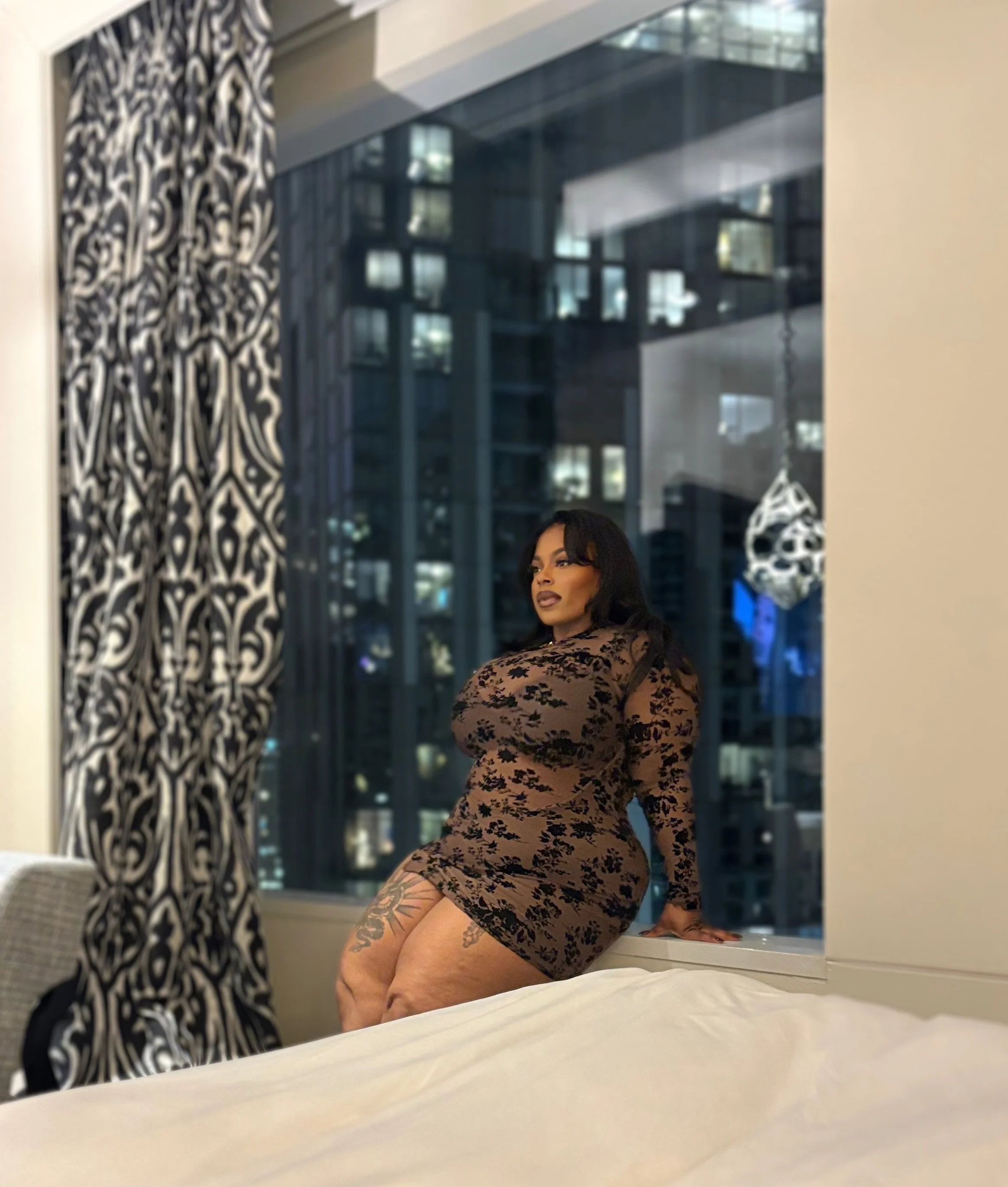 A woman with dark hair and tattoos, wearing a sheer black floral dress, sitting on a bed by a large window in a high-rise apartment at night, with city lights visible outside.