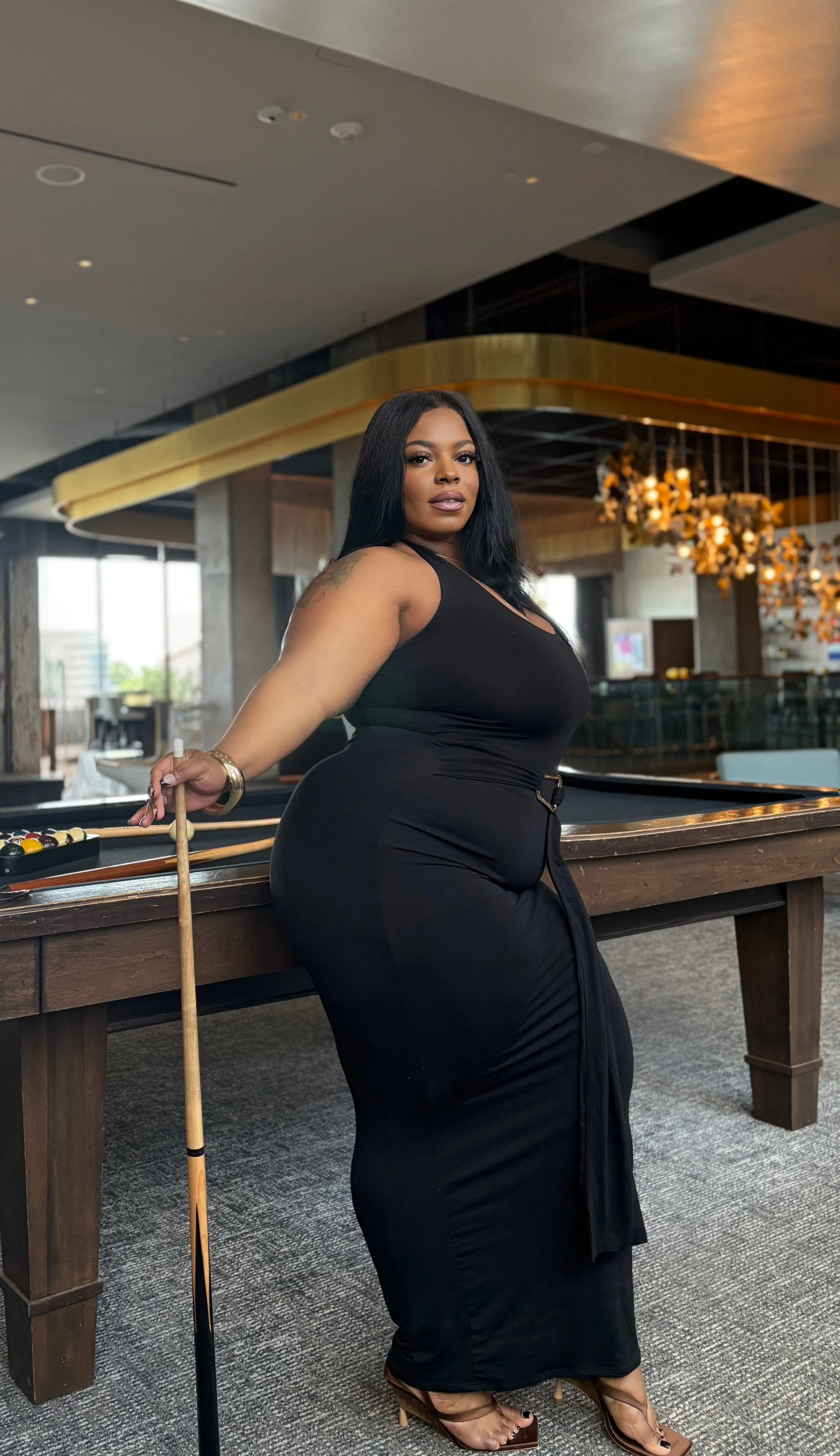 A woman in a black dress standing next to a pool table in a modern, upscale lounge or casino, holding a pool cue with a city view in the background.