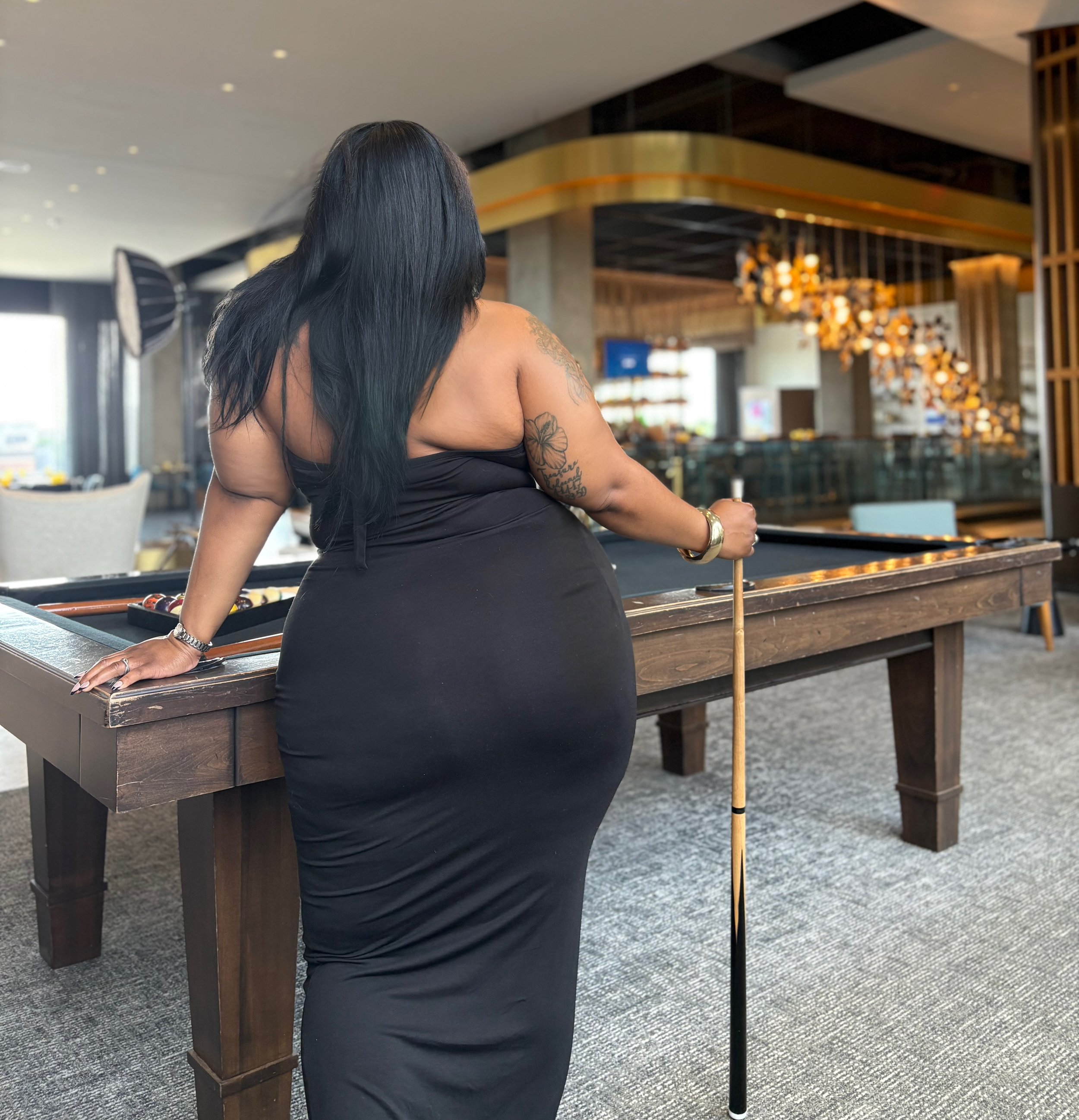 A woman with long black hair, tattoos on her arm, and a black dress, standing by a pool table in a modern lounge or hotel restaurant.