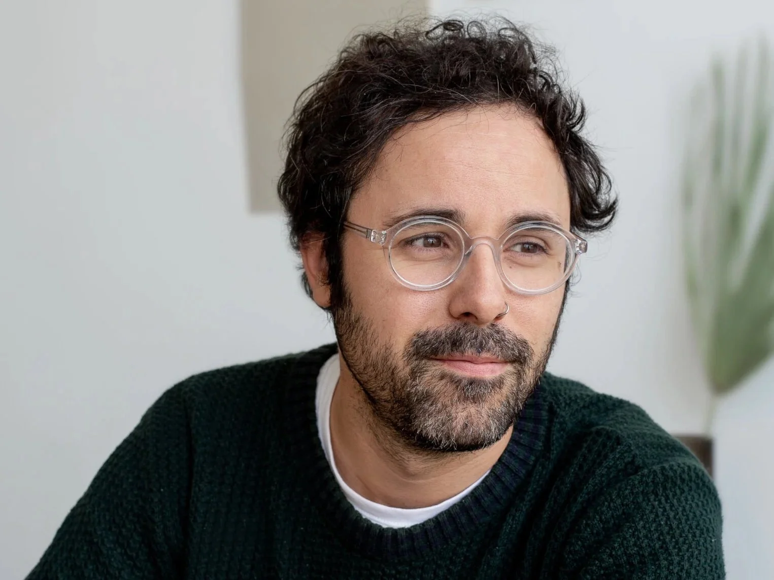 Portrait of a man with brown hair and a beard, wearing clear round glasses, a black sweater, and a white undershirt, looking thoughtfully to the side indoors.