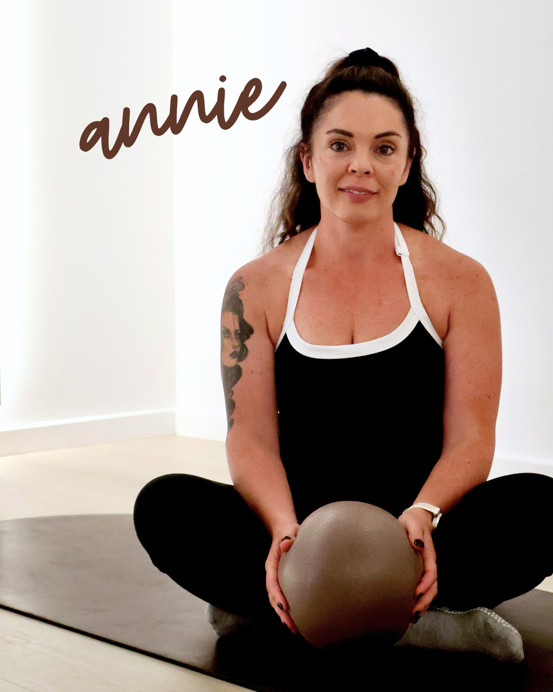 A woman with dark, curly hair tied up in a bun, sitting cross-legged on a black yoga mat, holding a stability ball, wearing a black and white athletic top, with a tattoo on her left arm, in a bright, minimal indoor space.