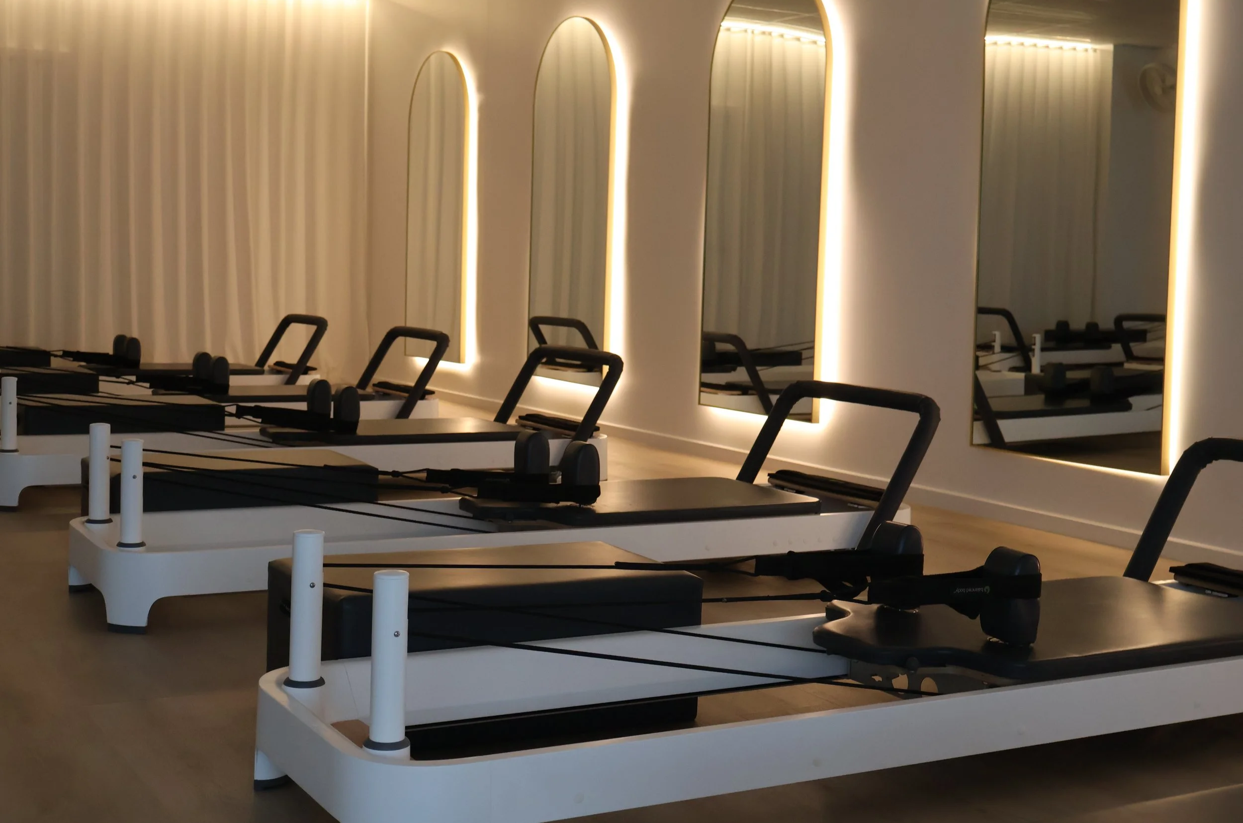 A Pilates studio with several reformer machines lined up in front of a wall with tall, illuminated mirrors and white curtains.
