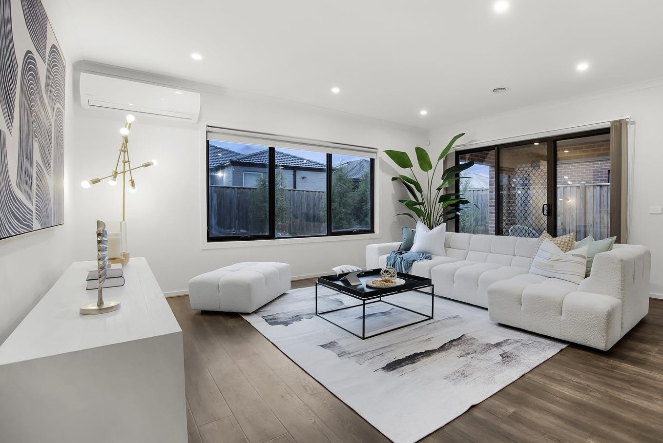 Minimalist home office with white desk, modern chair, and white blinds. Lamp and books on desk. Large abstract art and plant beside dark cabinet. Hardwood floor.