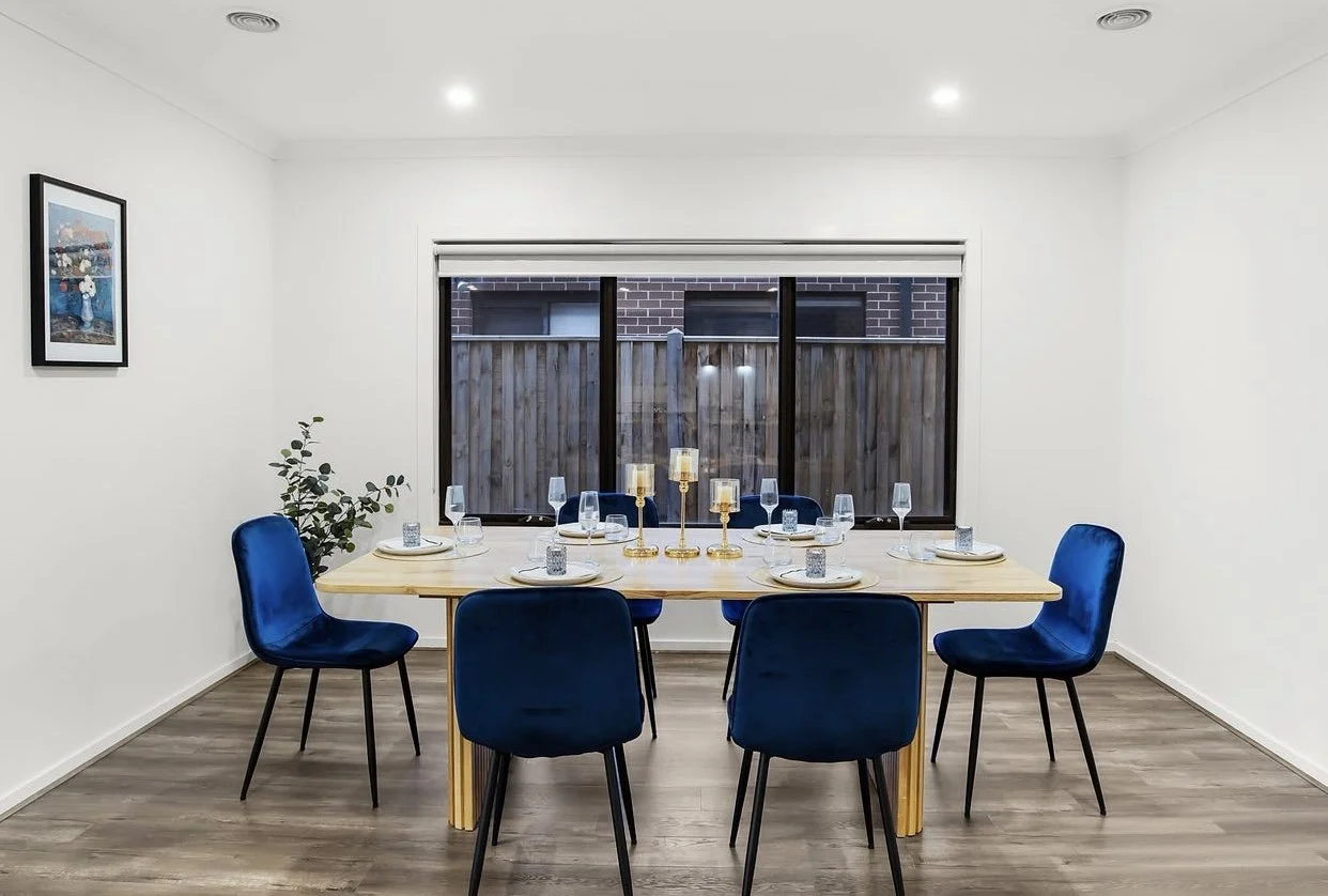 Modern dining room with a wooden table set for six, featuring blue upholstered chairs, and decorative pendant lights above. Glass sliding doors and large windows provide natural light. A staircase with wooden rails is visible on the left.