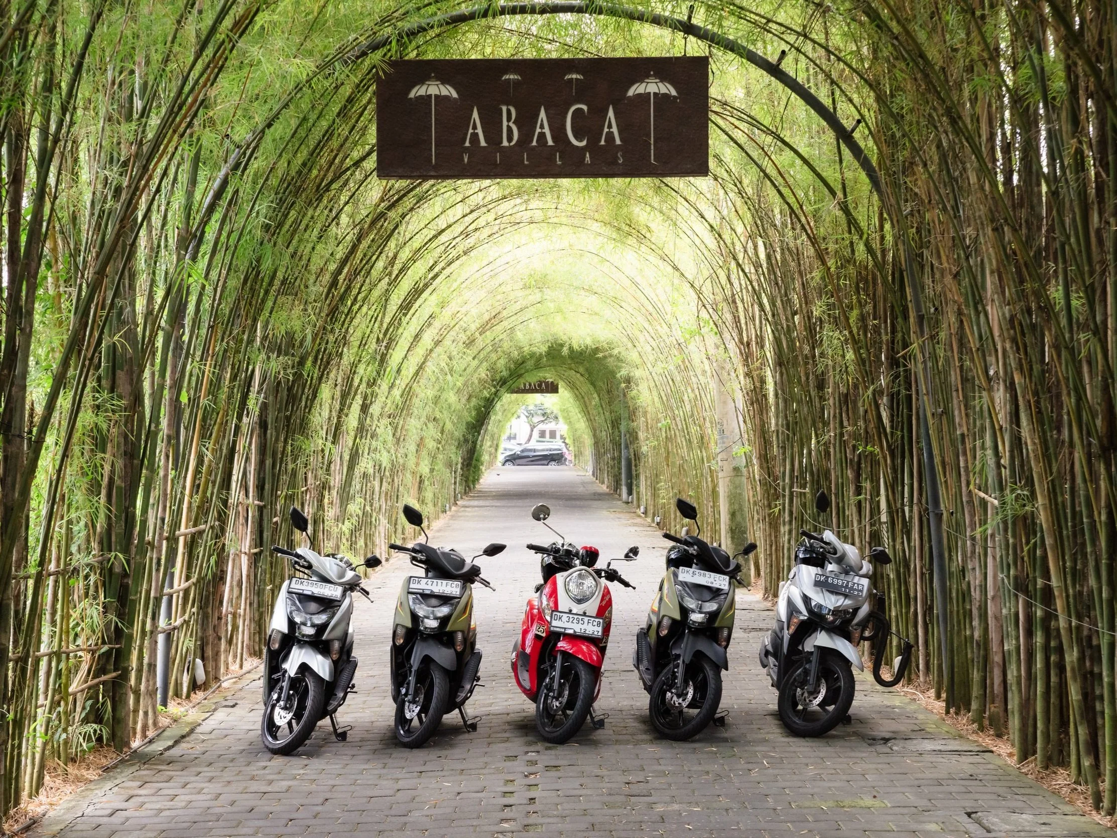 Five motorcycles parked under a bamboo archway in a tunnel-like pathway leading through a green bamboo forest. A sign hanging above reads "ABACA Villlas."