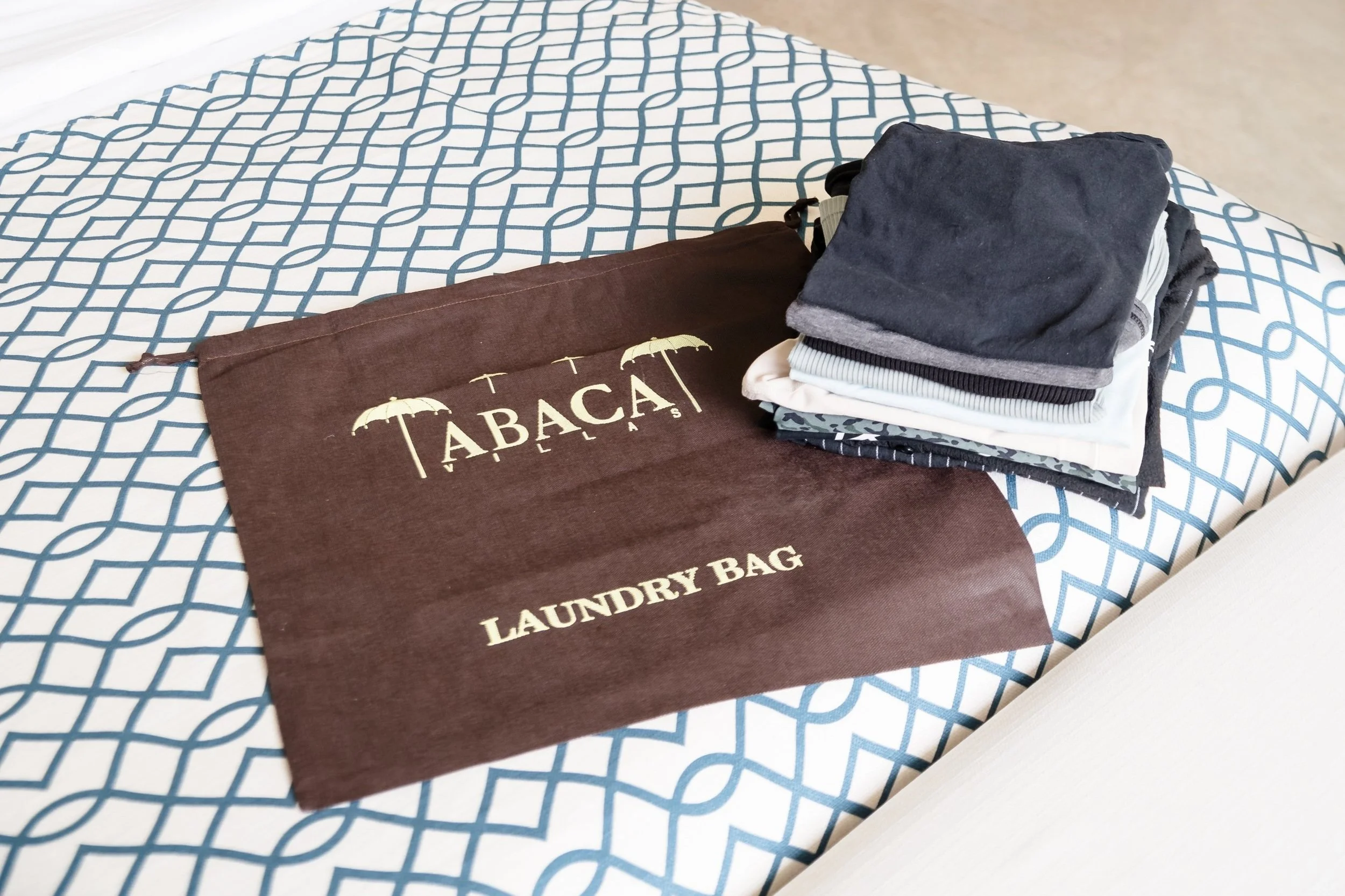A brown laundry bag with the words 'Tabacá Laundry Bag' and umbrella graphic on it, placed on a bed with a blue and white geometric patterned bedsheet. Next to the bag, there is a neatly folded stack of various cloth items, including dark, light, and patterned fabrics.