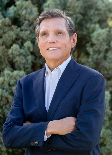 Un hombre de mediana edad sonriente, vestido con un traje azul marino y camisa blanca, de pie al aire libre con follaje verde al fondo.