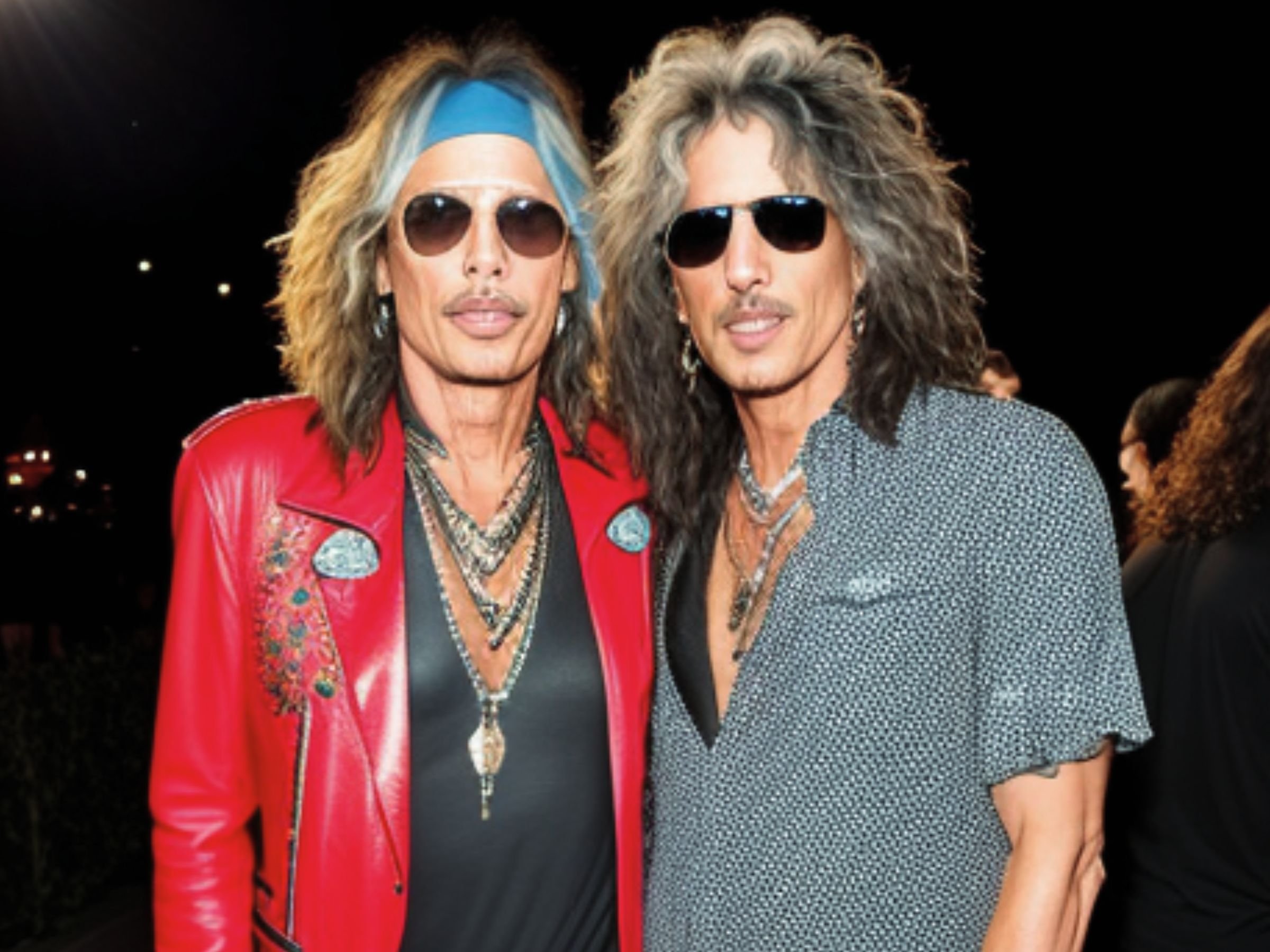 A photograph of legendary rockers, Steven Tyler and Joe Perry, sitkcing their tongues out at a red carpet event at night.