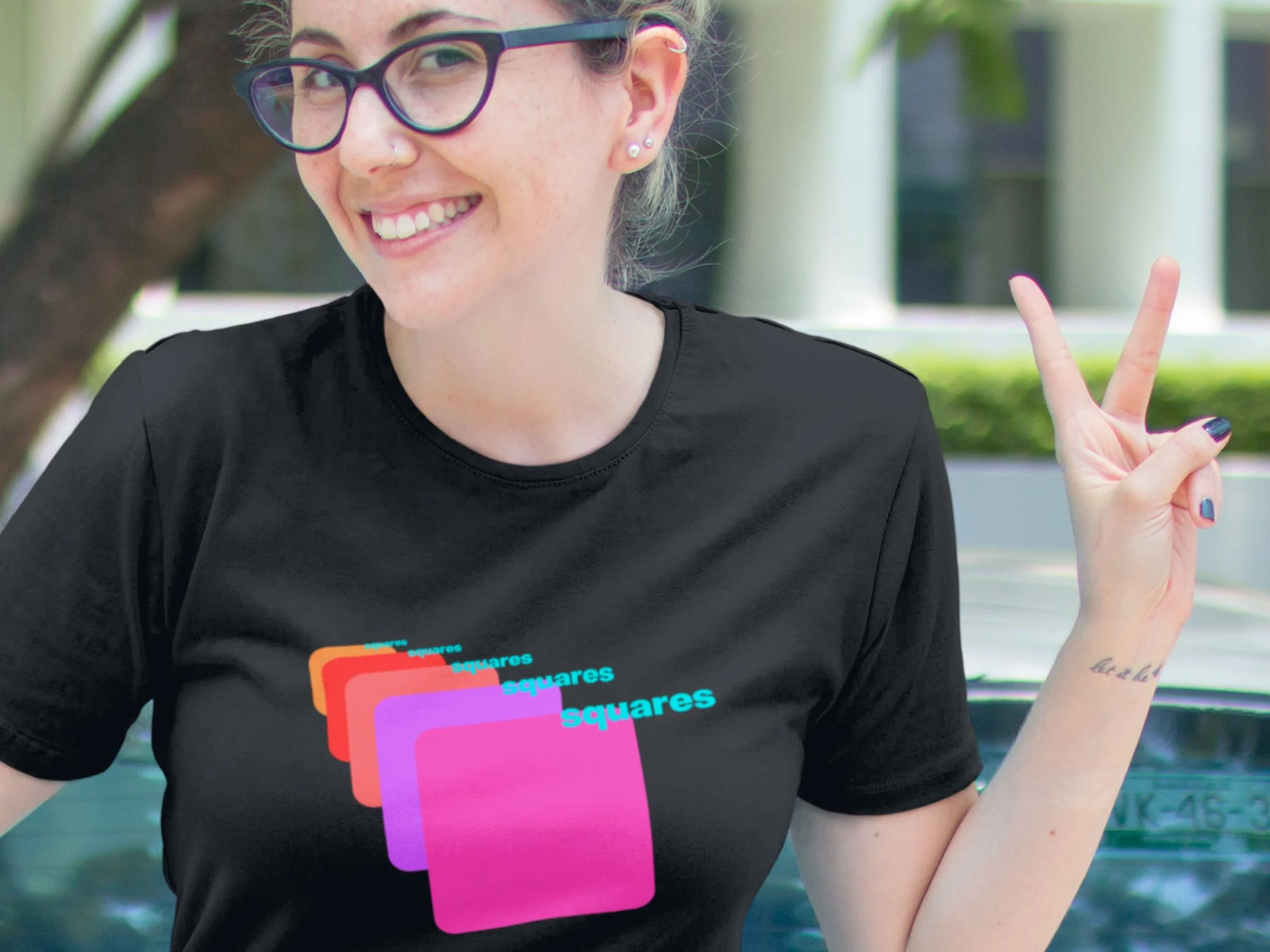 A young woman wearing glasses and flashing the peace sign is wearing a black t- shirt from Don Q's Private Hams featuring multi-colored squares receeding across the shirt.