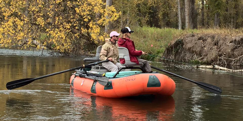 A man and a woman fish in an orange boat as the float down a river surrounded by autumn leaves