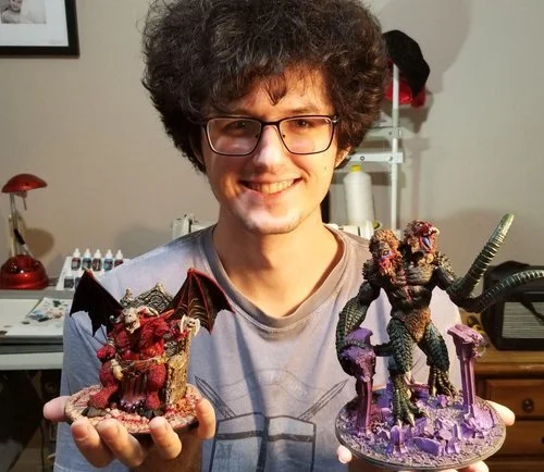A young man with curly hair and glasses smiling, holding two detailed fantasy-themed figurines, one in each hand, in a room with a desk, lamp, and shelves in the background.