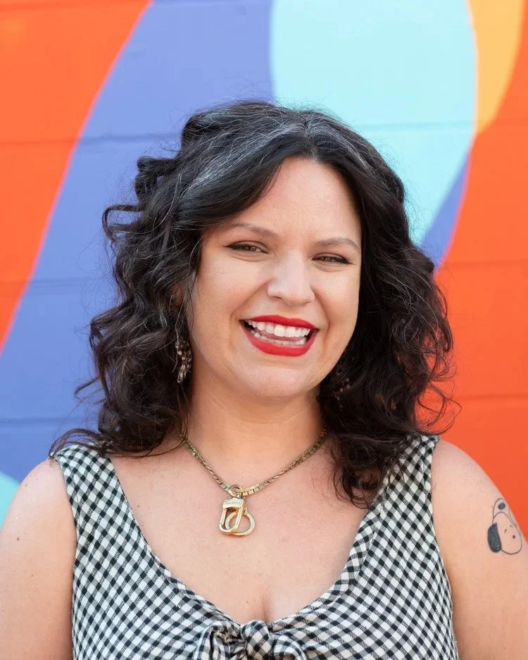 A woman with curly dark hair smiling in front of a colorful rainbow umbrella background.