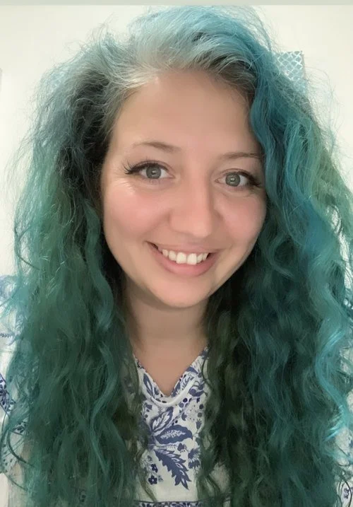 A young woman with long, curly hair that is a mix of grey and teal, smiling at the camera. She is wearing a white top with a blue floral pattern, and the background is indoors with light-colored walls.
