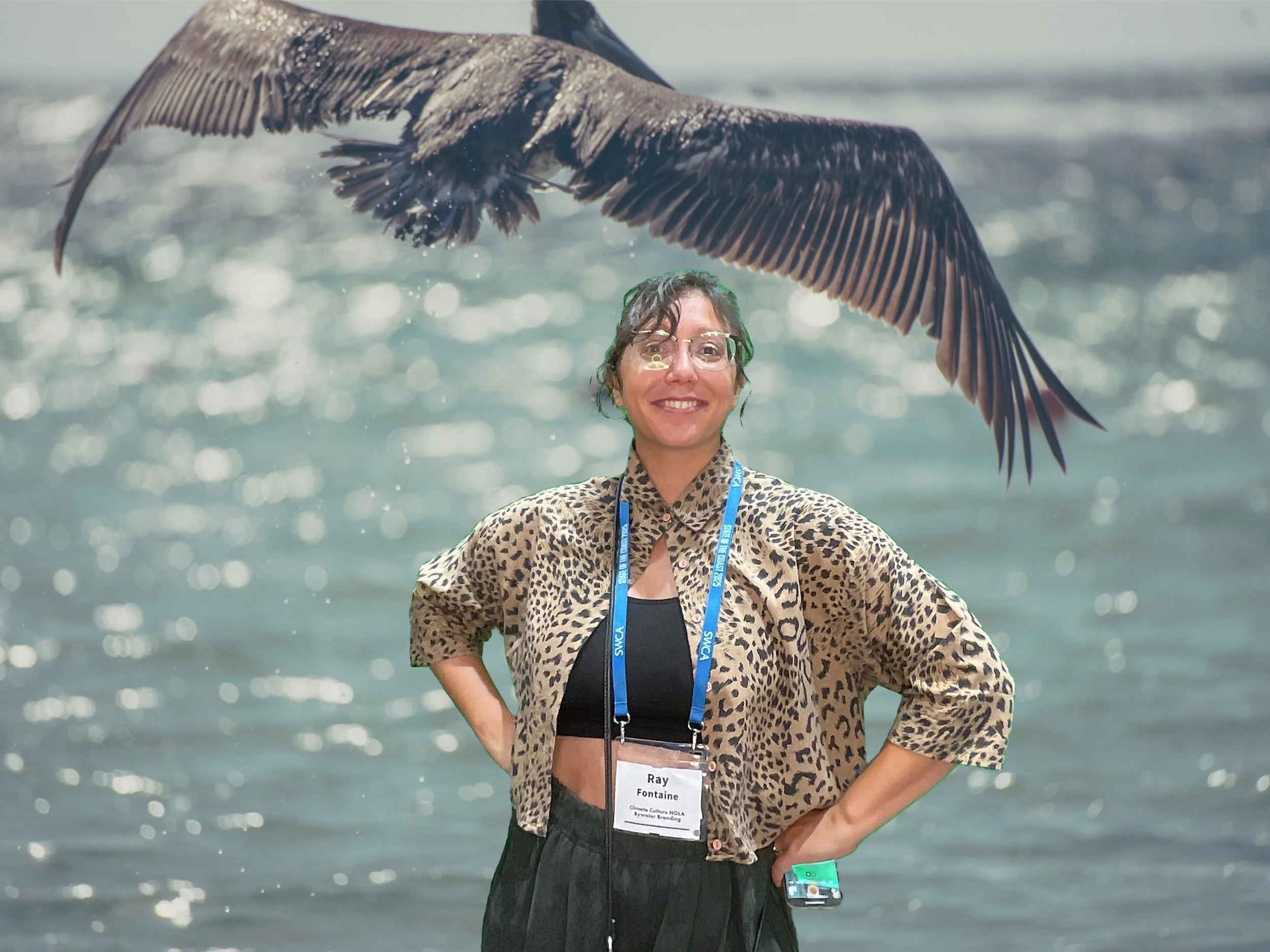 A woman with glasses and short curly hair smiling at the camera, standing in front of a body of water, wearing a leopard print jacket, black top, and a lanyard with a badge, with a large bird flying overhead.