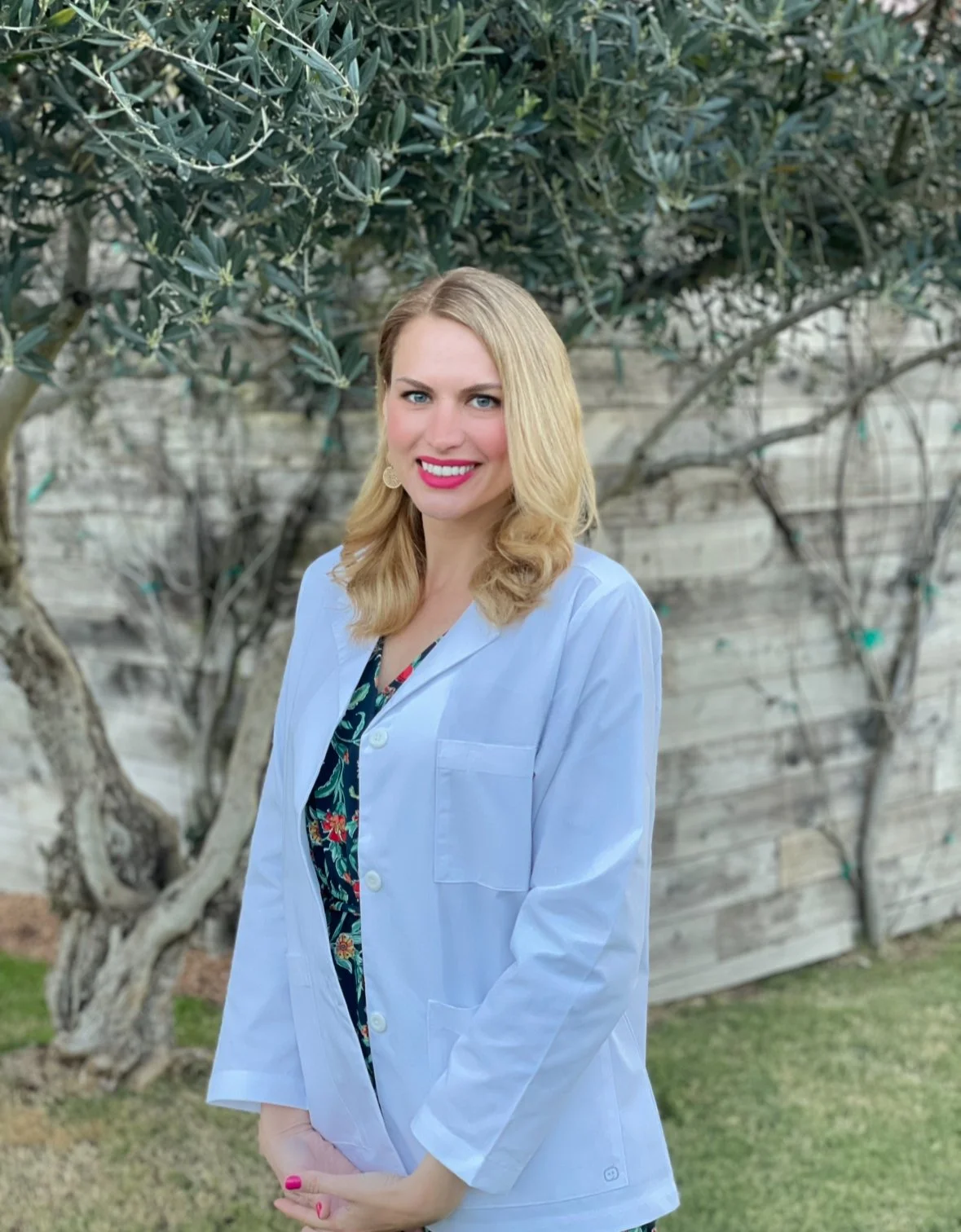 A woman with blonde hair, wearing a white lab coat, standing outdoors in front of a tree and a wooden fence, smiling.