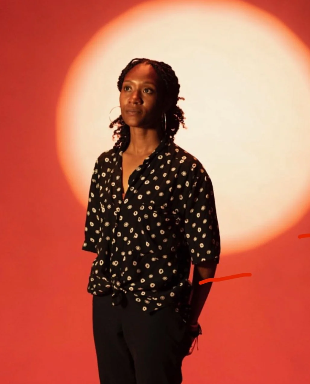 A woman with braided hair, wearing hoop earrings and a black shirt with white polka dots, standing against a vibrant orange background with a large glowing white circle behind her.