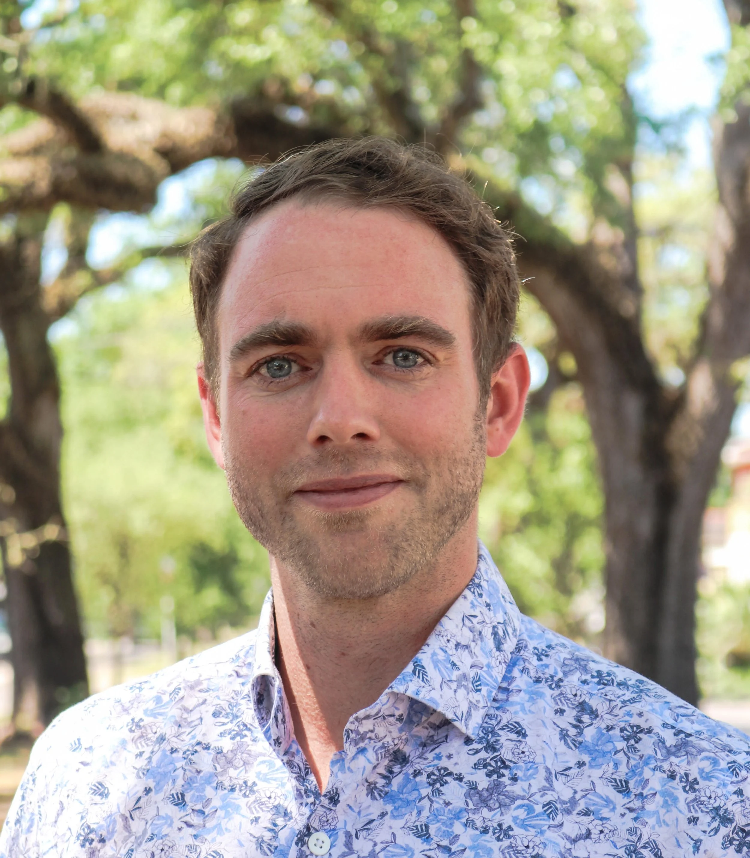 A young man with light skin, brown hair, and blue eyes standing outdoors in front of green trees, wearing a white shirt with a blue floral pattern.