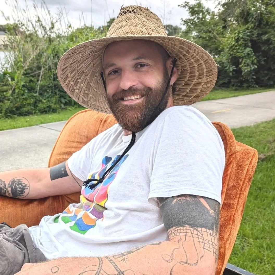 A smiling man with a beard, wearing a straw hat and a white T-shirt, sitting on a rust-colored outdoor chair in a grassy area with trees in the background.