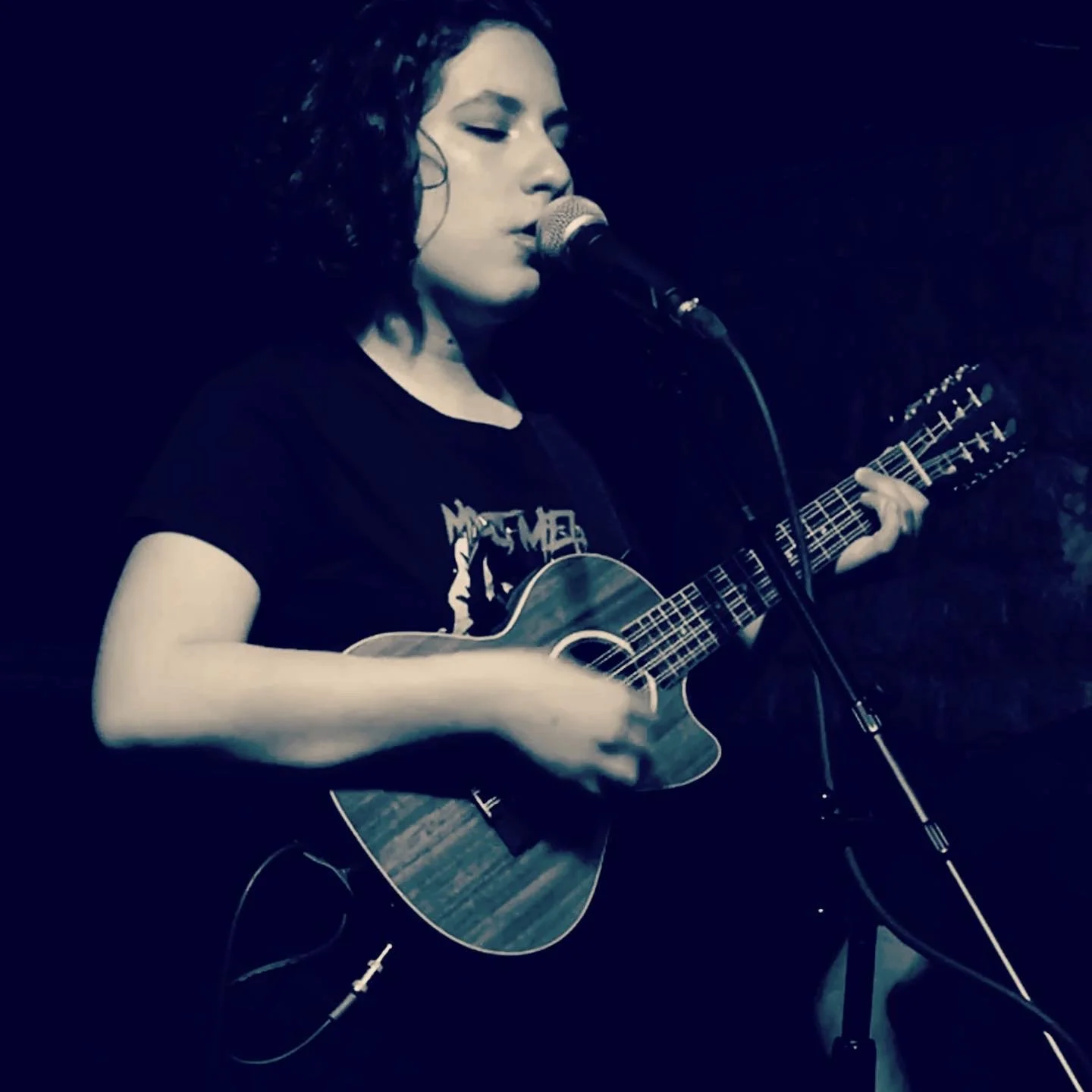 A woman with curly hair singing into a microphone while playing an acoustic guitar on stage.