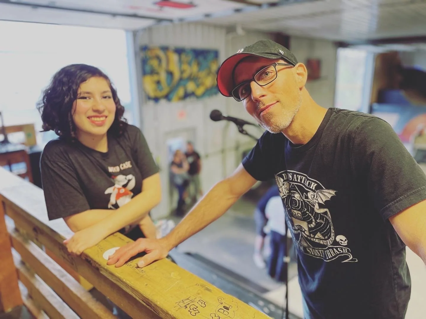 A woman with curly black hair and a man with glasses and a baseball cap smiling at the camera inside a skate shop or skate park, with a wooden railing and skateboards in the background.