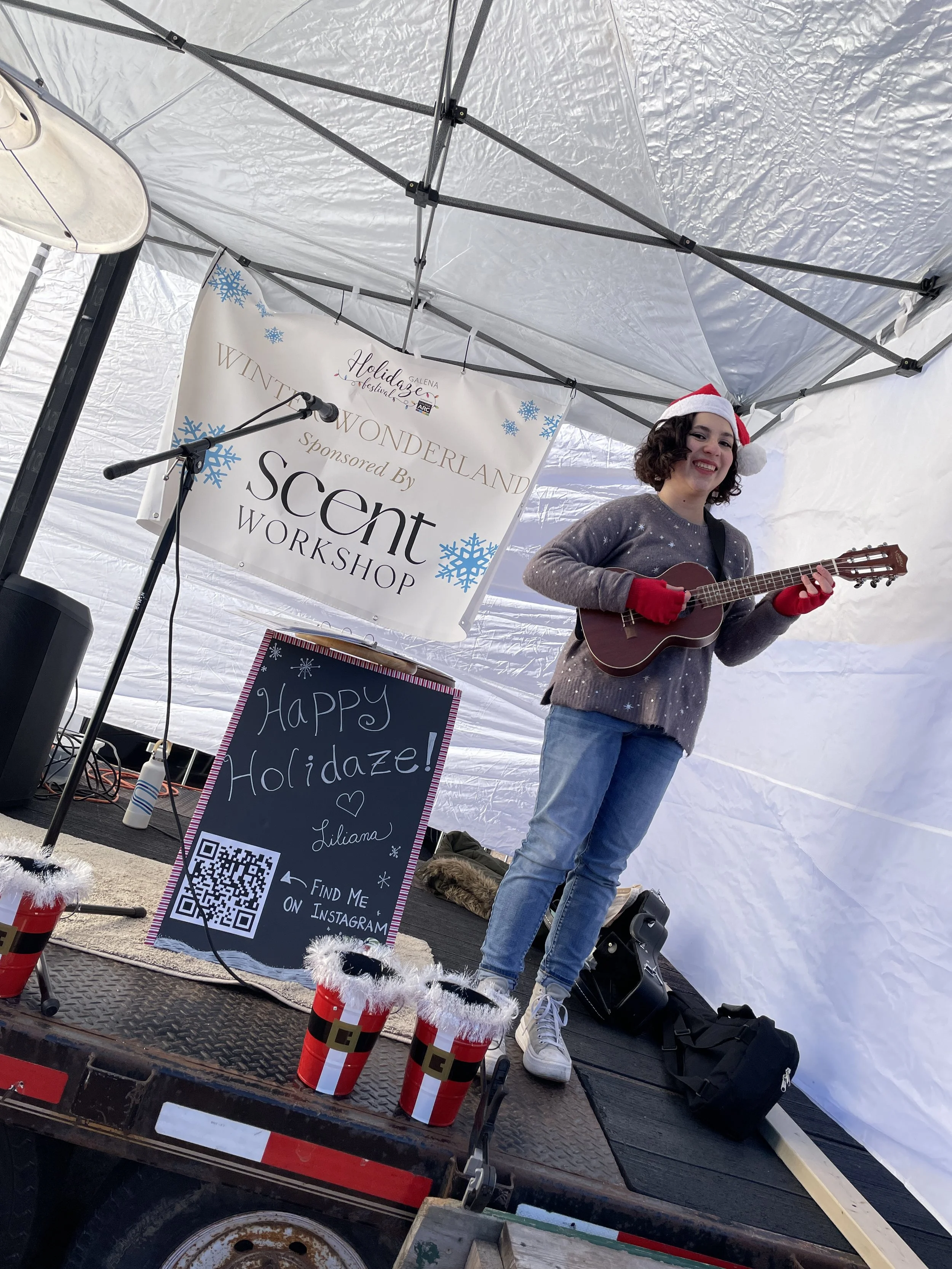A woman wearing a Santa hat, red gloves, and a gray sweater with white details, is playing a ukulele on a small outdoor stage at a holiday event. There is a banner behind her that reads 'Winter Wonderland Sponsored By Scent Workshop' and a chalkboard