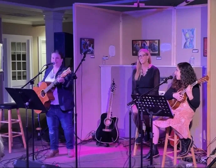 Three women performing live music on a small stage, with two women playing guitars and singing, and one woman singing without an instrument. Colorful stage lighting creates a vibrant atmosphere. Artwork hangs on the walls behind them.