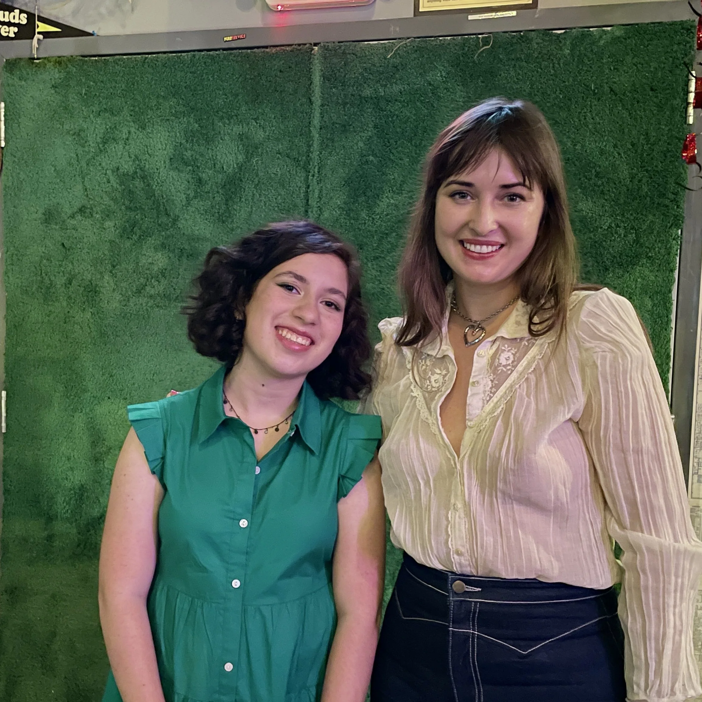 Two women smiling and standing close together in front of a green backdrop, one wearing a teal dress with ruffled sleeves and the other wearing a cream-colored blouse with vertical stripes.