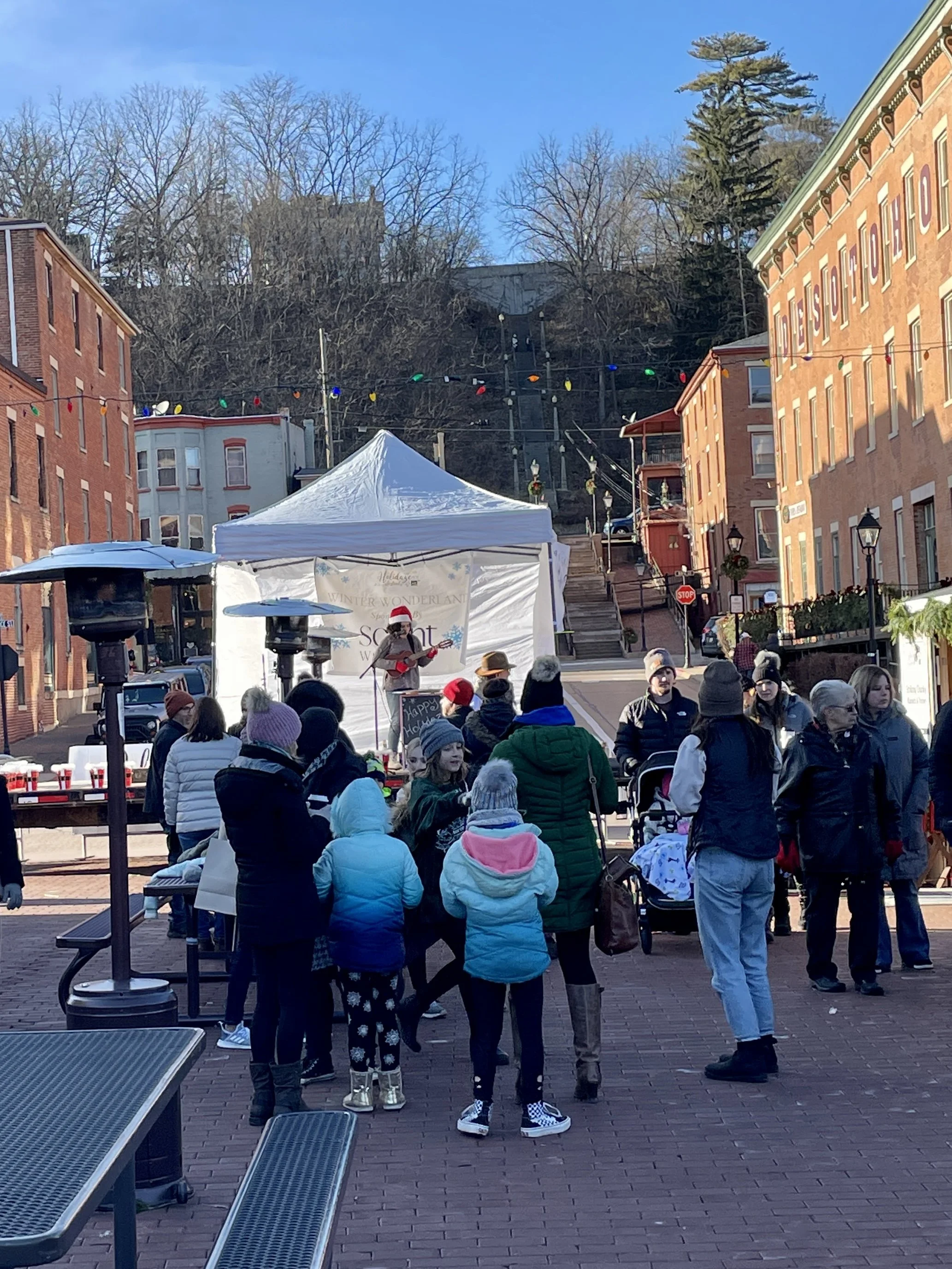 People gathered outdoors on a brick-paved street with some wearing winter clothing, watching a performer on a small stage under a white tent, with festive holiday decorations and colorful string lights overhead.
