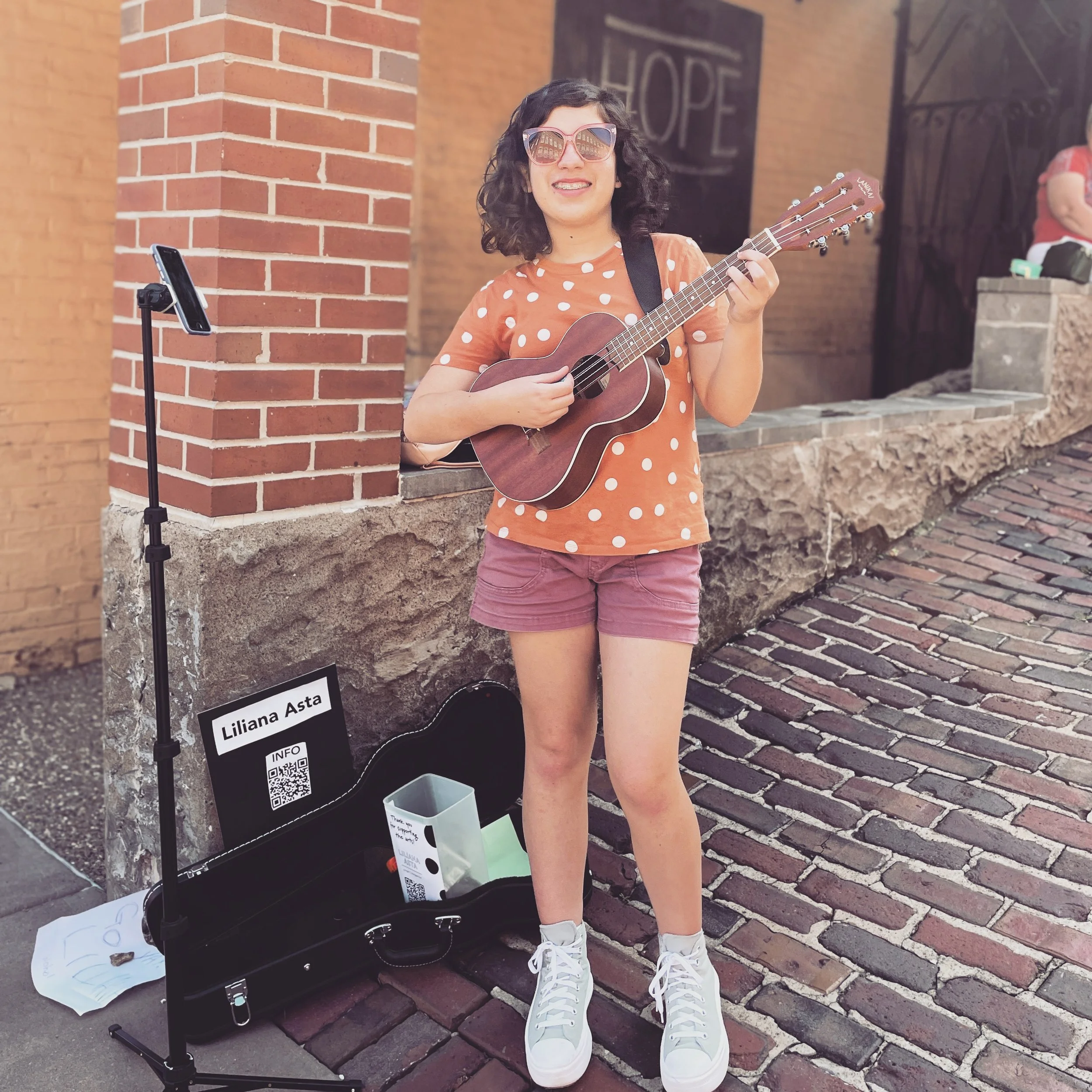 A woman wearing sunglasses, an orange polka-dot shirt, and pink shorts, is standing on a brick sidewalk, playing a ukulele. She is smiling, and there is a small open case on the ground with a sign that says 'Liliana Asta' and a QR code. A smartphone 