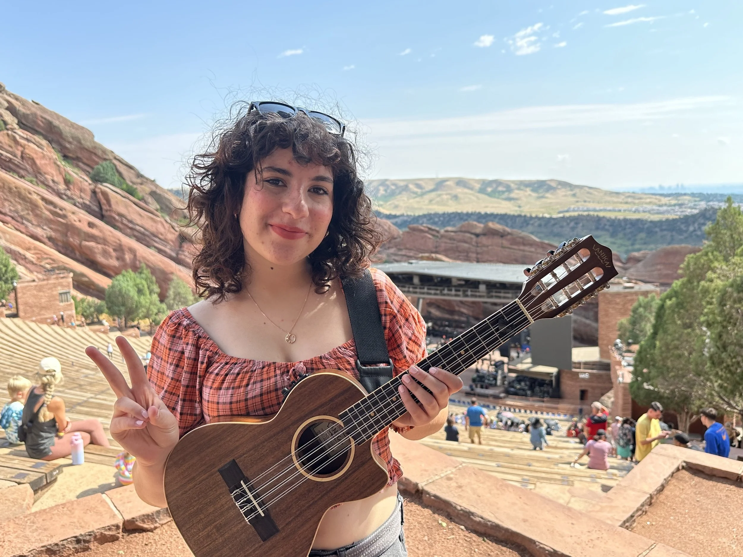 Young woman with curly hair wearing a plaid top holding a ukulele and making a peace sign outdoors in a rocky amphitheater with people and trees in the background.