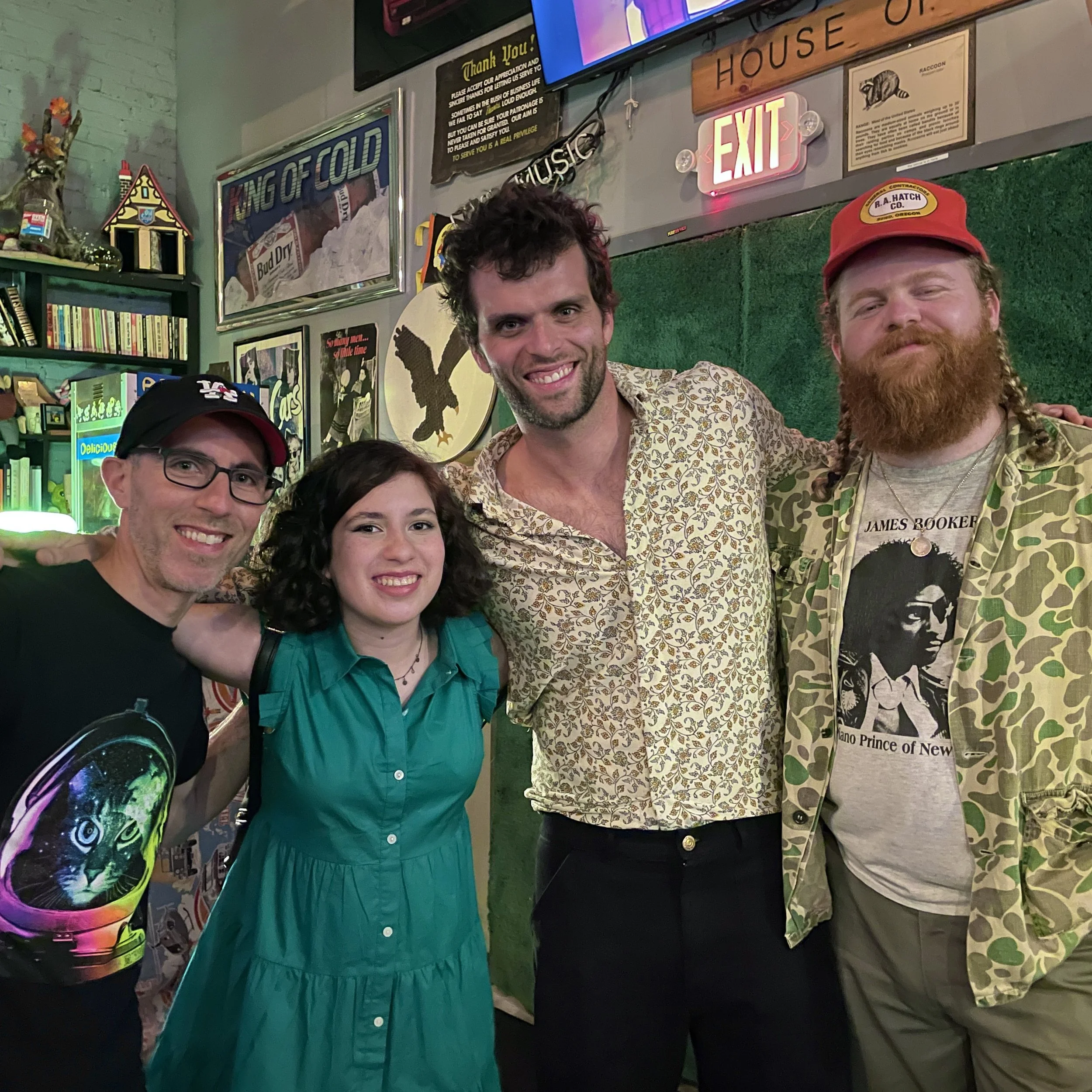 Four people standing together in a bar or restaurant, smiling at the camera. The setting has various signs and decorations on the wall behind them, including a neon exit sign, a framed picture, and a sign that says 'King of Cold.' The group includes 