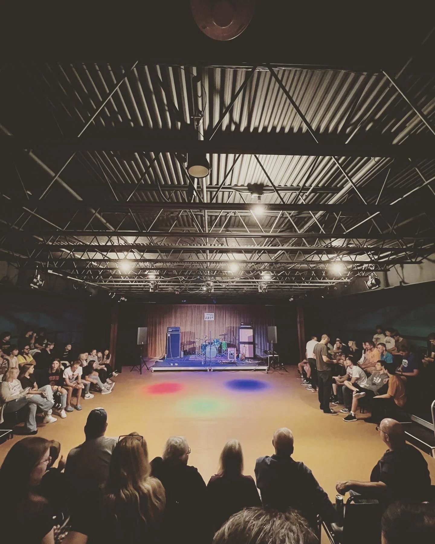 Audience waiting for a music performance on stage in a venue with a high ceiling and colorful spotlights on the dance floor.