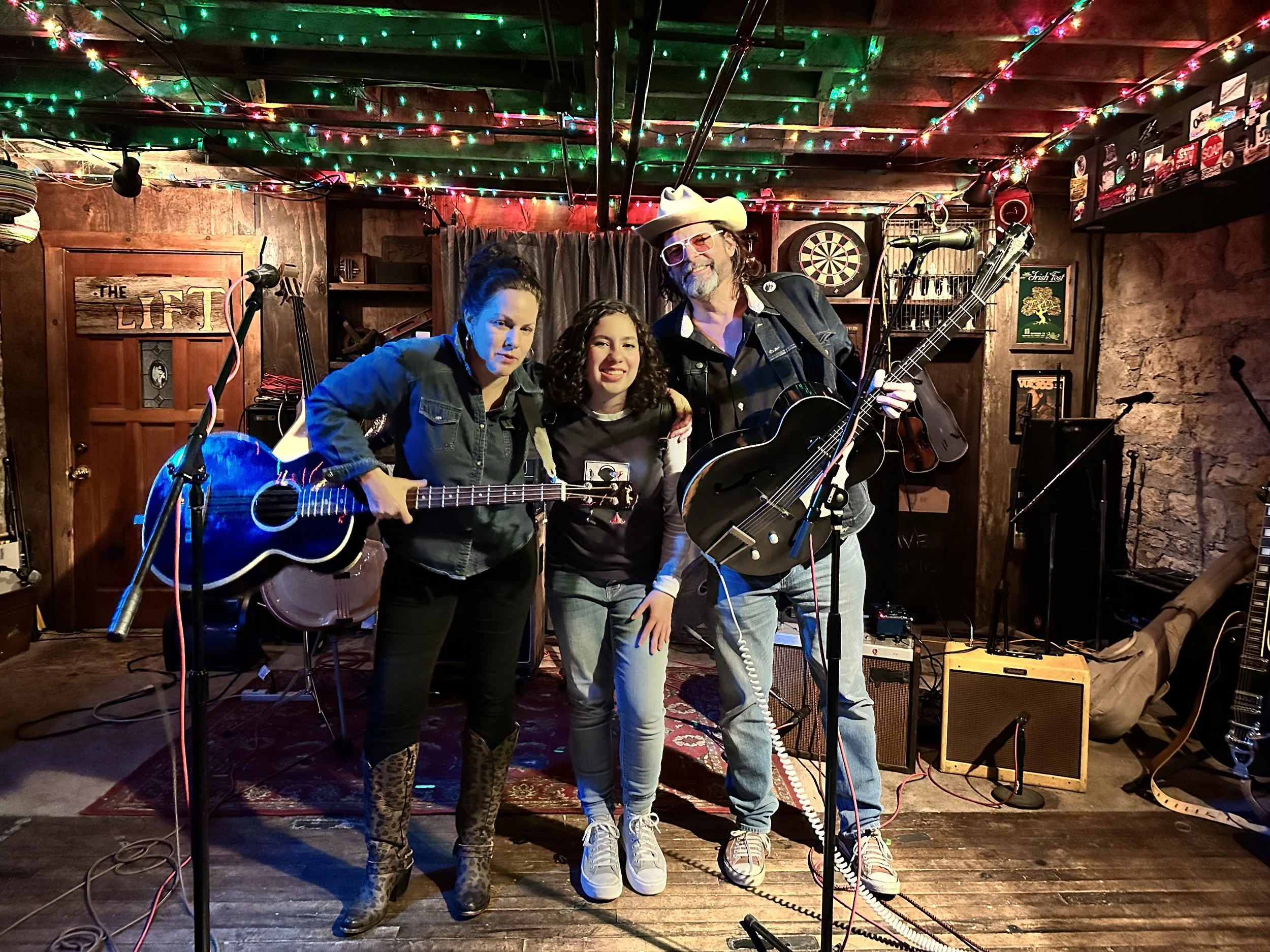 Three musicians standing together on a stage with musical instruments and equipment in a cozy, decorated venue with colorful string lights overhead.