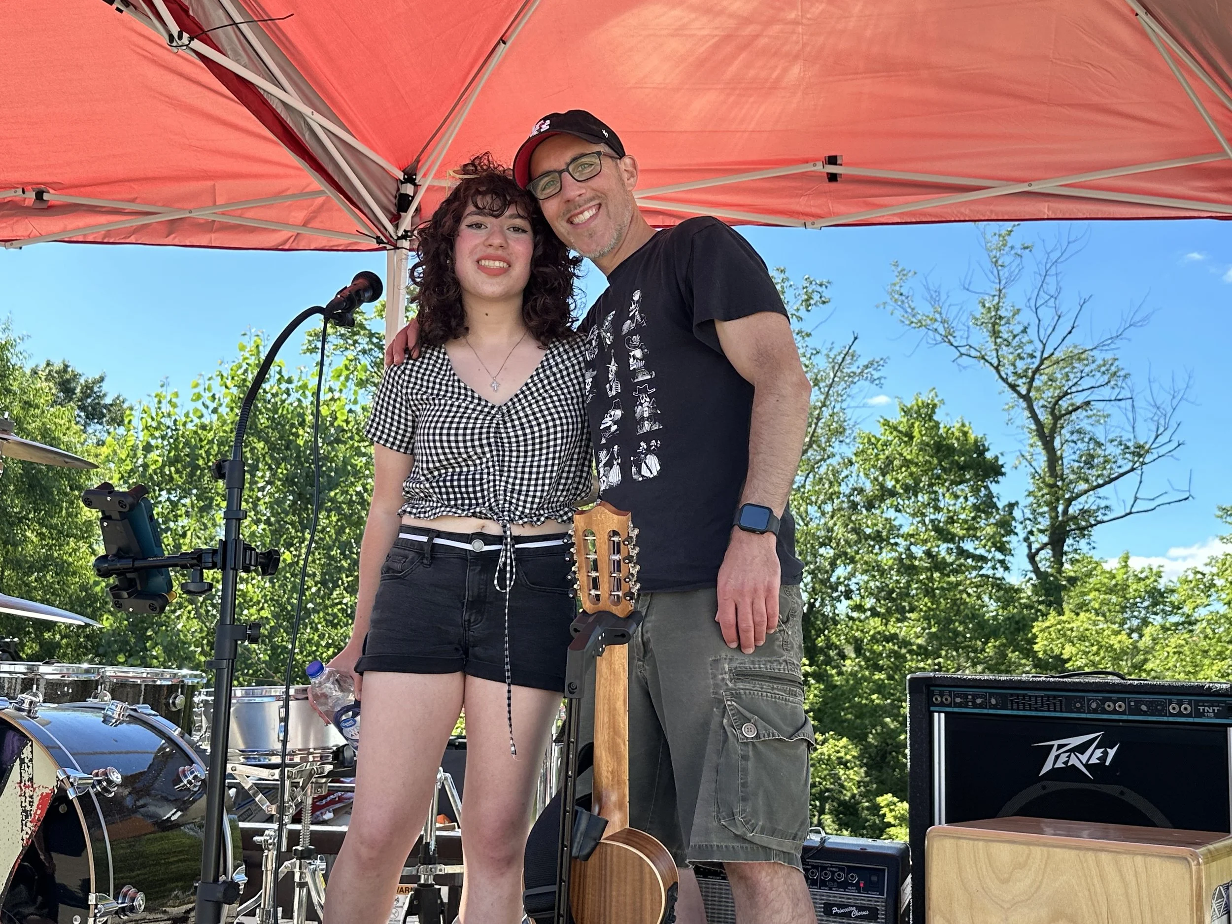 A young woman and a man standing outdoors under a red canopy, smiling for a photo. The woman is wearing a black and white checkered top and black shorts, holding a water bottle. The man is wearing glasses, a black t-shirt, gray shorts, and a smartwat