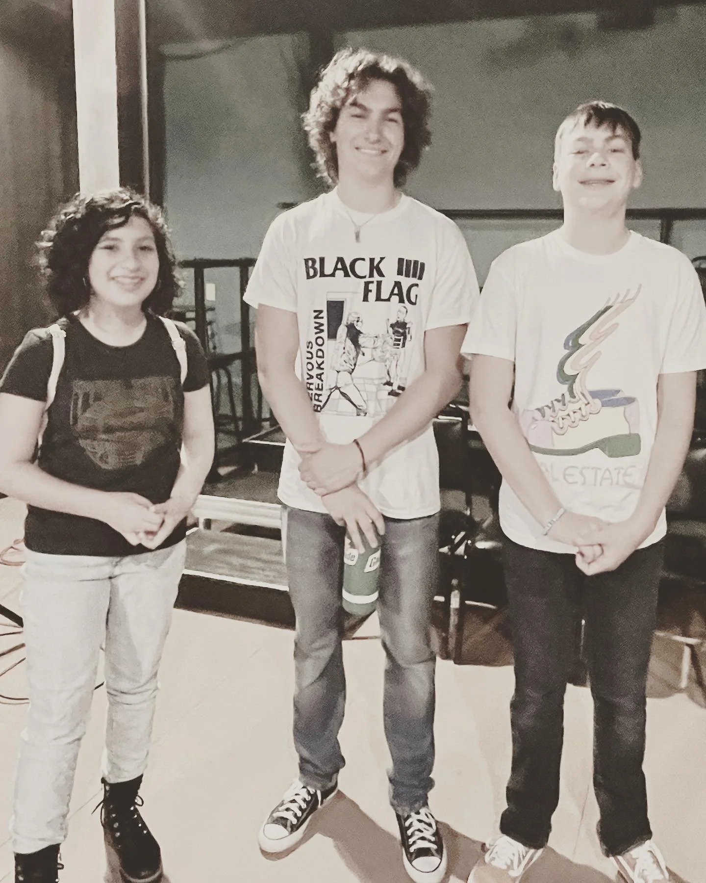 Three teenagers standing indoors, smiling at the camera. The girl on the left has curly dark hair, wearing a black T-shirt and light-colored pants with black boots. The boy in the middle has shoulder-length curly hair, wearing a white graphic T-shirt