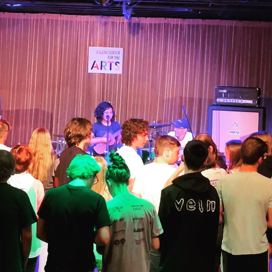 A live music performance at the Galena Center for the Arts with a female singer, guitarist, and band members on stage, and an audience watching in front.