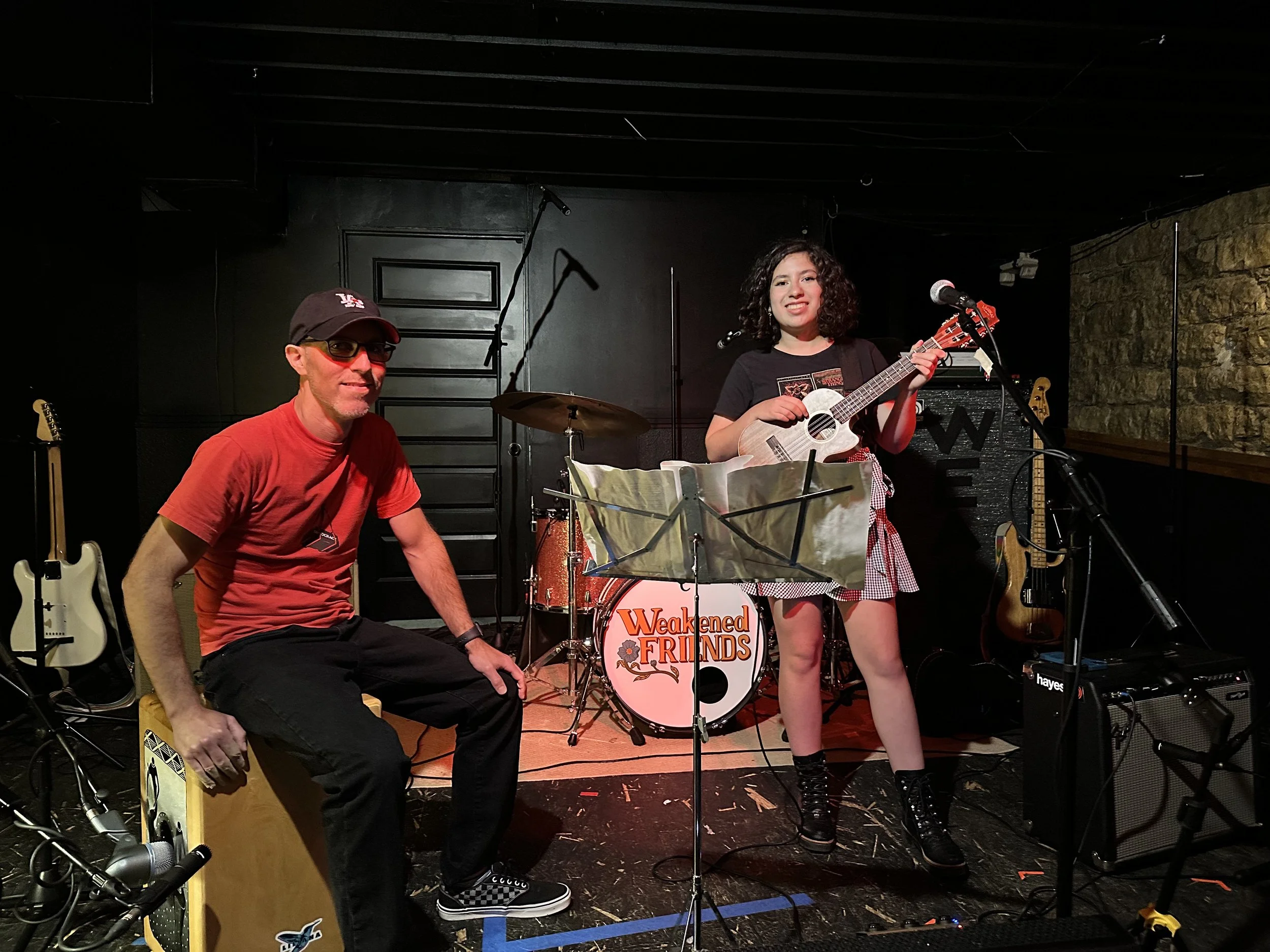 Two musicians performing on stage, one woman playing guitar and singing, one man sitting on a cajón drum, with drums, guitars, and amplifiers in the background.