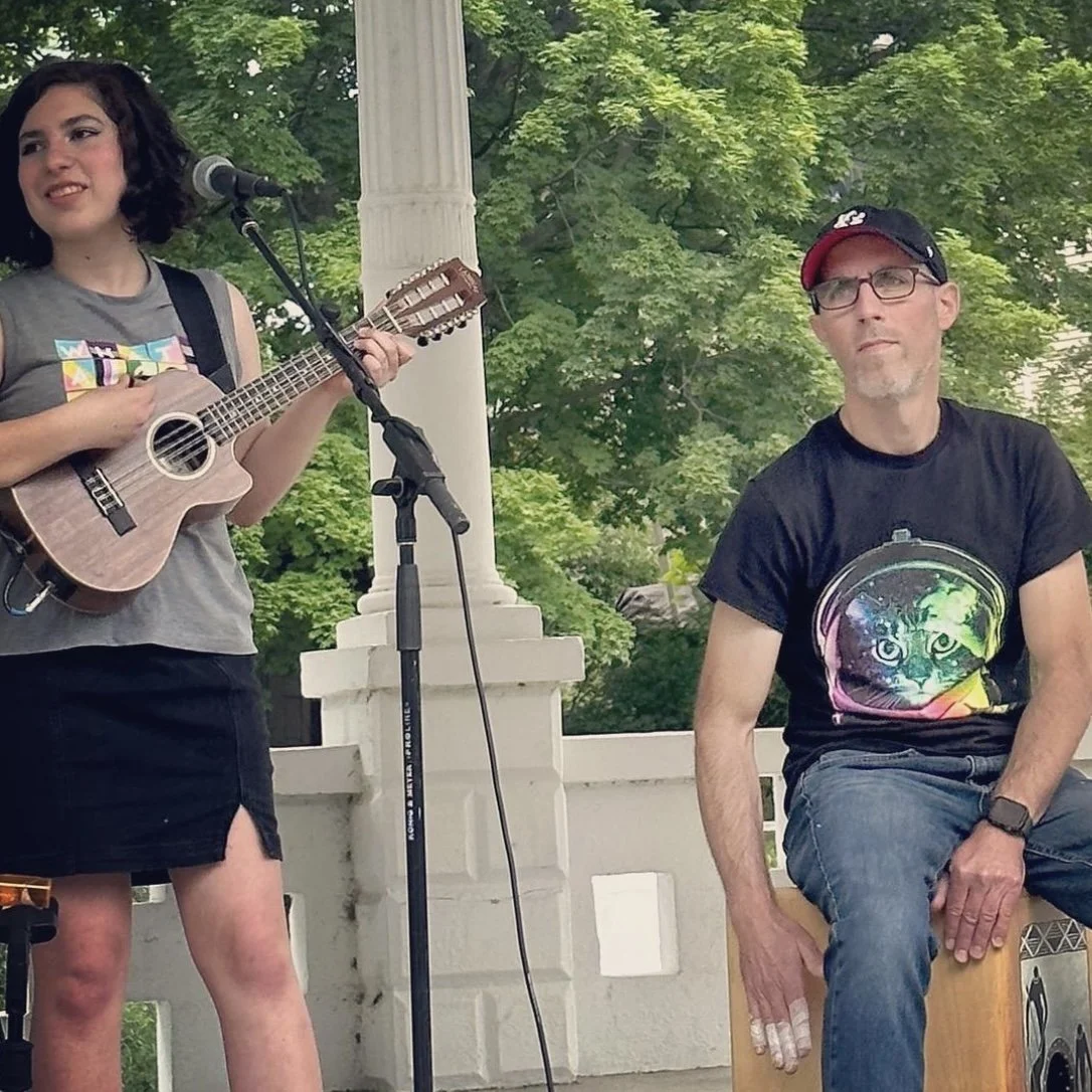 Young woman singing and playing ukulele, man seated on cajón, outdoor setting with trees and white railing.