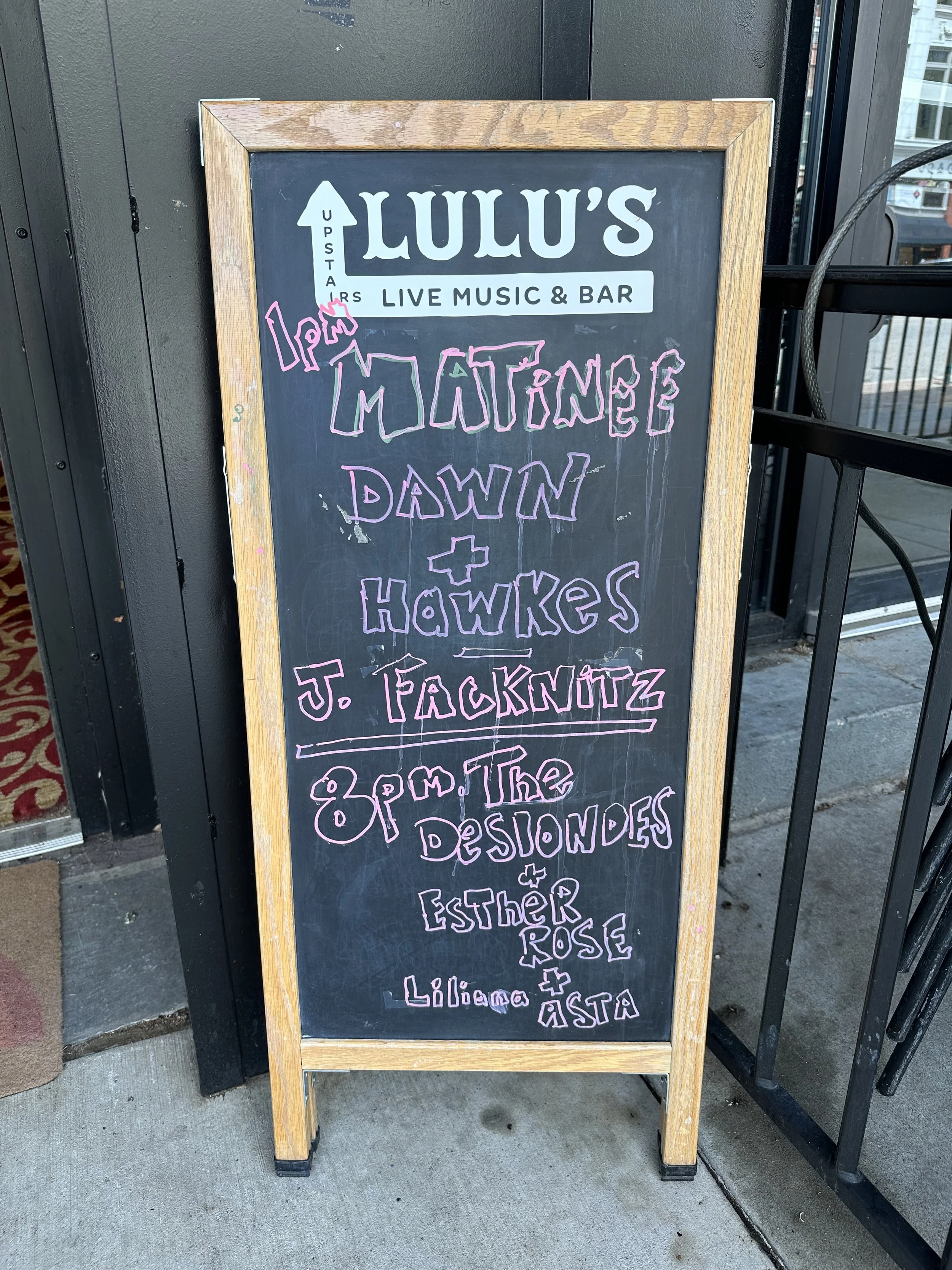 A blackboard outside Lulu's Live Music & Bar advertising a music event with bands Matinee Dawns, Hawks, J. Facknitz, The Desondes, Esther Rose, Liliano, and Åsta, scheduled for 8 p.m.
