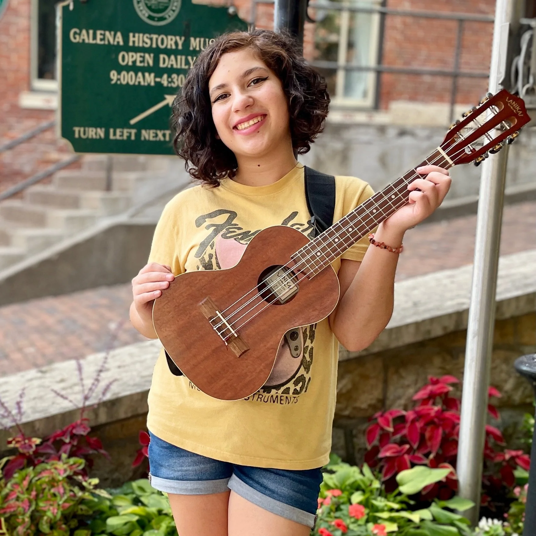 Young woman with curly dark hair smiling and holding a ukulele outside in front of a sign for Galena History Museum.