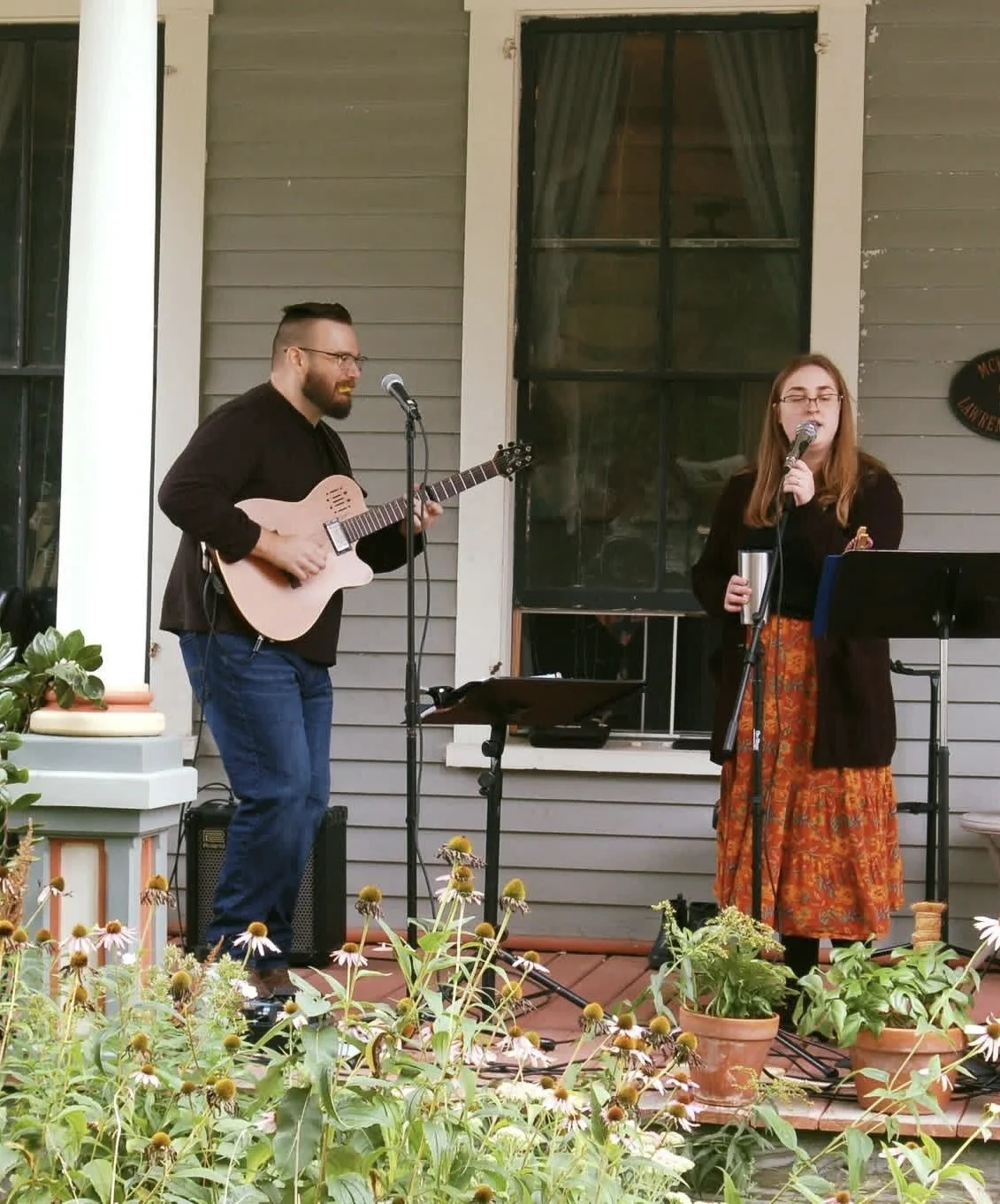 Two people performing music outdoors in front of a house. The man on the left plays an acoustic guitar and sings into a microphone, while the woman on the right sings into a microphone and holds a cup. There are plants and flowers in pots in the foreground.