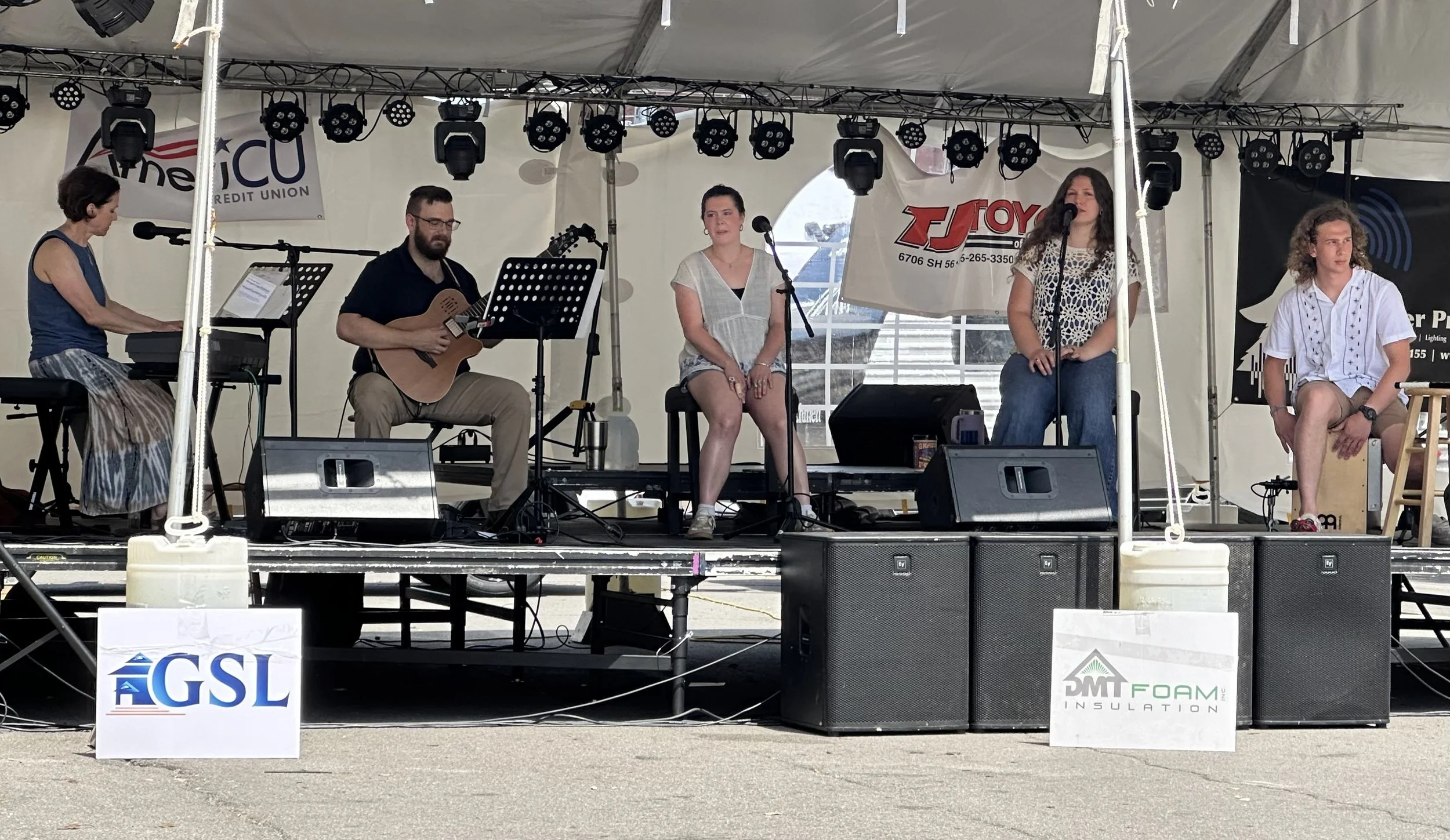 A band performing on a stage under a tent at an outdoor event, with five musicians playing instruments and singing.