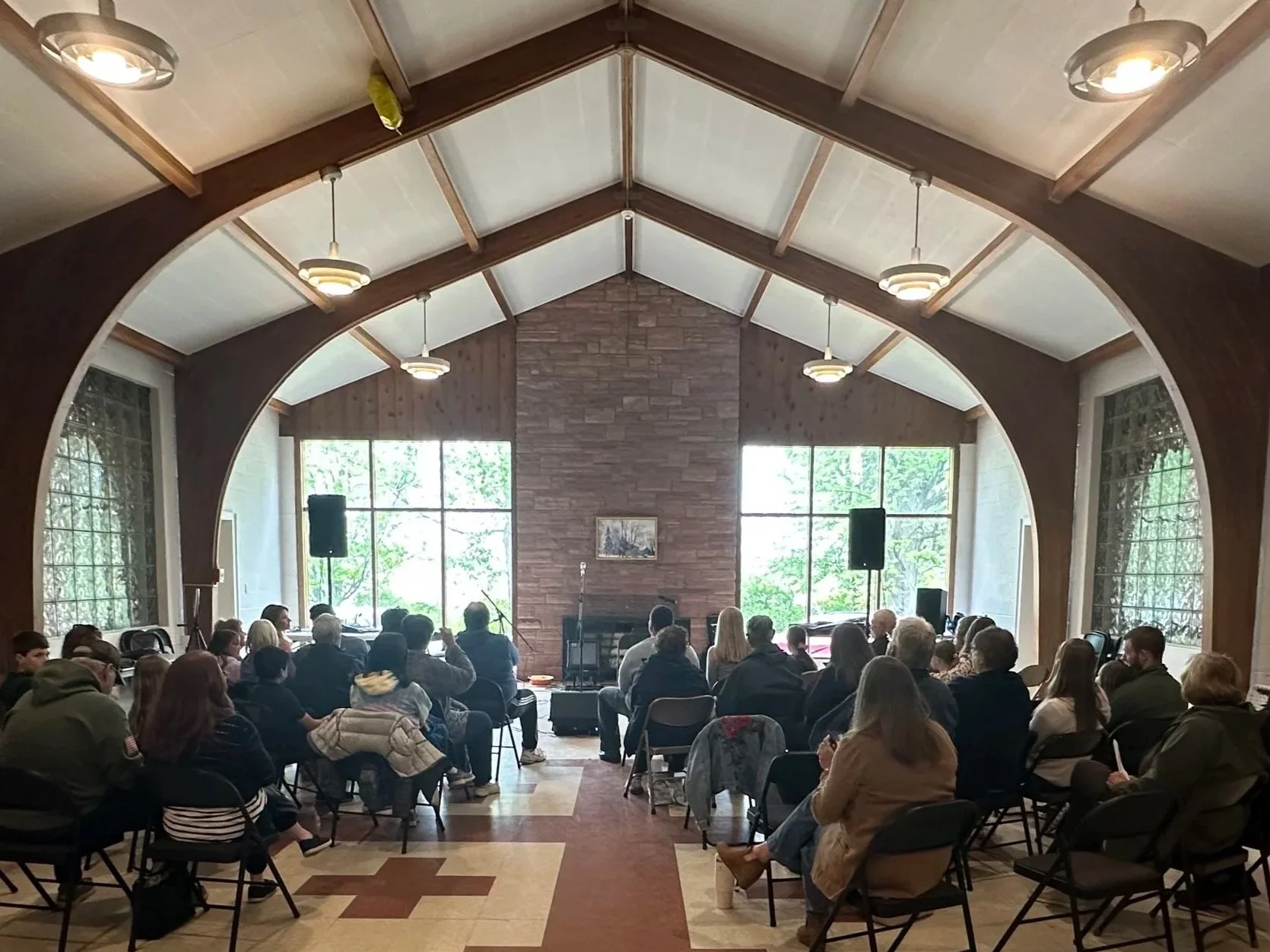 A room with a high ceiling, large windows, and wood and brick walls, filled with people sitting on chairs, attending a presentation or performance at the front with microphones, speakers, and a painting on the wall.