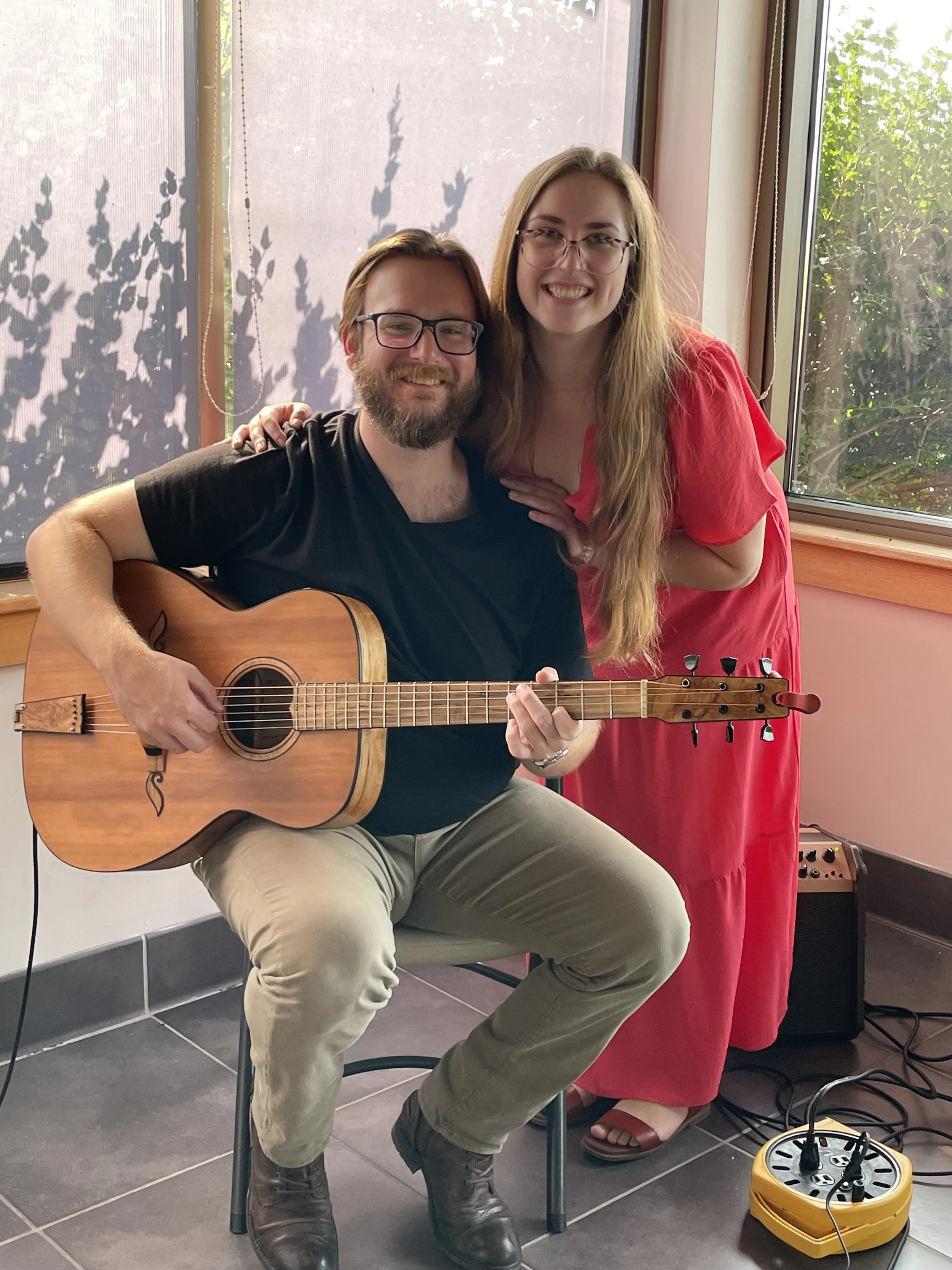 A man sitting on a chair playing an acoustic guitar, smiling, while a woman in a pink dress stands next to him, smiling and resting her hand on his shoulder, inside a room with large windows and greenery outside.
