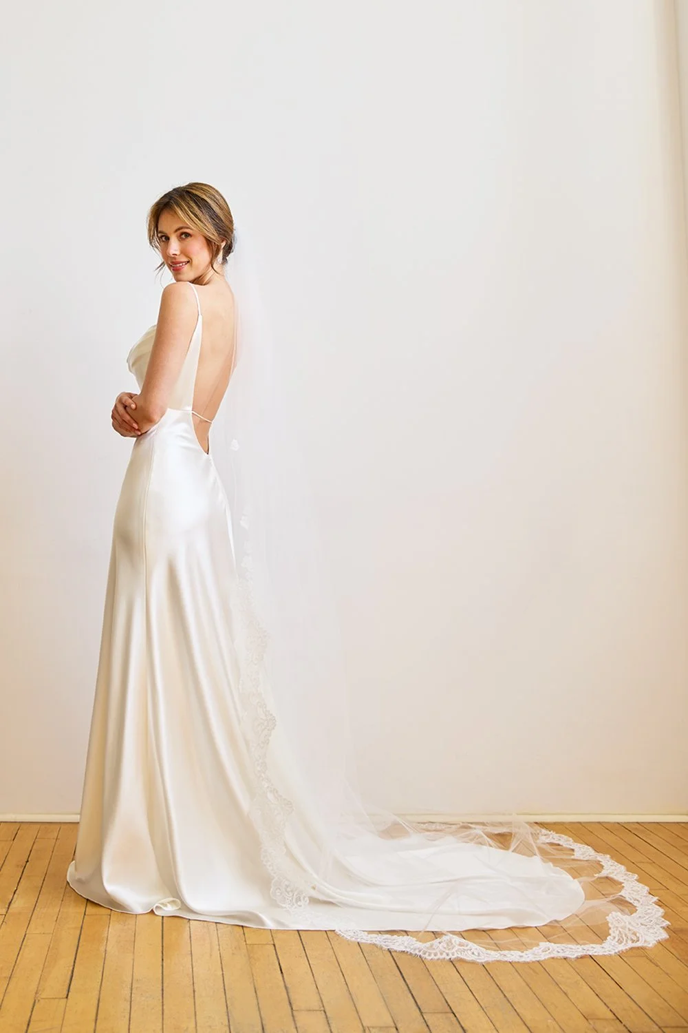A woman in a white satin wedding dress with a train and lace accents, standing on a wooden floor next to a white wall.