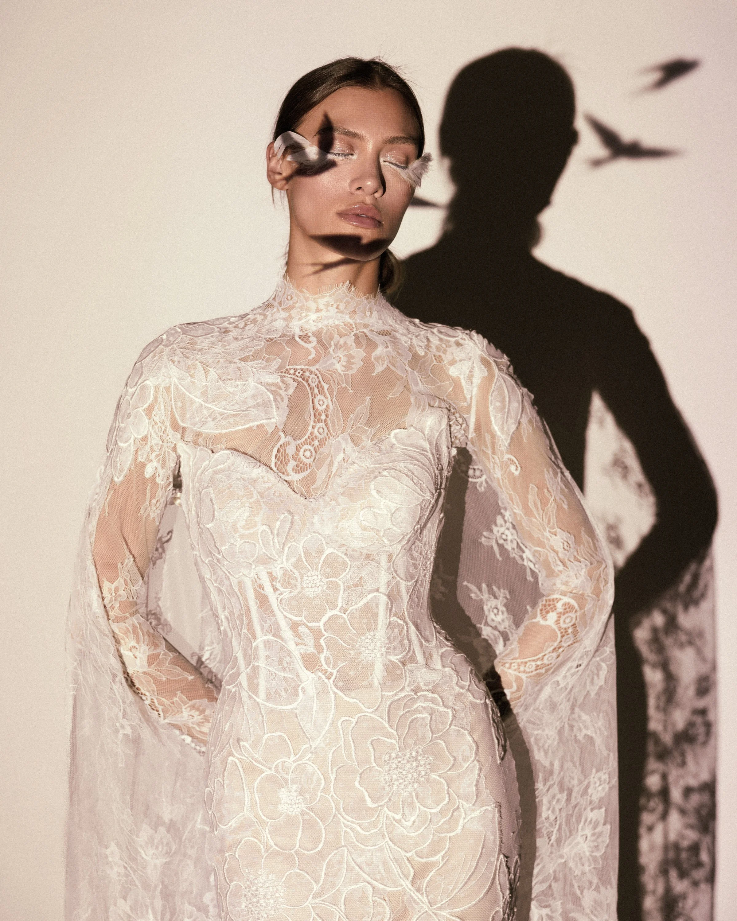 Gemy Maalouf at New York Bridal Makrket A woman with closed eyes wearing a lace dress, with a shadow and bird shadows cast on the wall behind her.