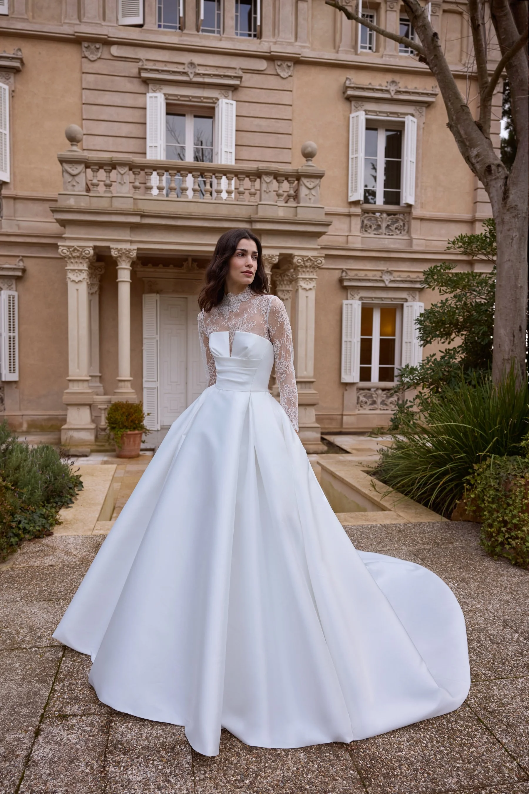 Jesus Peiro at New York Bridal Market.  A woman in a white wedding dress with lace sleeves standing outside in front of a large, elegant building with multiple windows and white shutters.