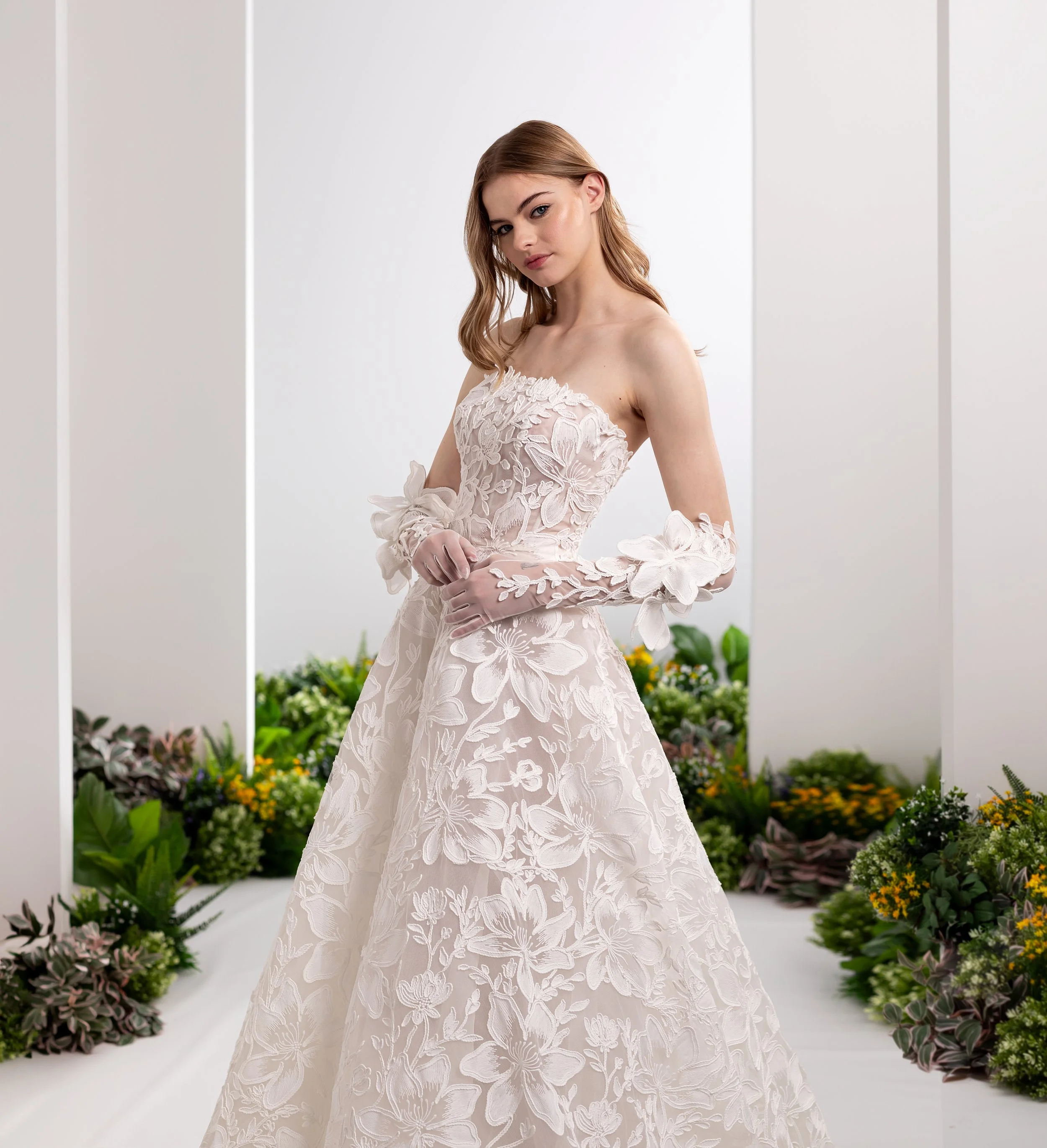 Valentini Spose at New York Bridal Market.  .A young woman in a white floral dress standing in front of a backdrop with greenery and flowers, posing for a photo.