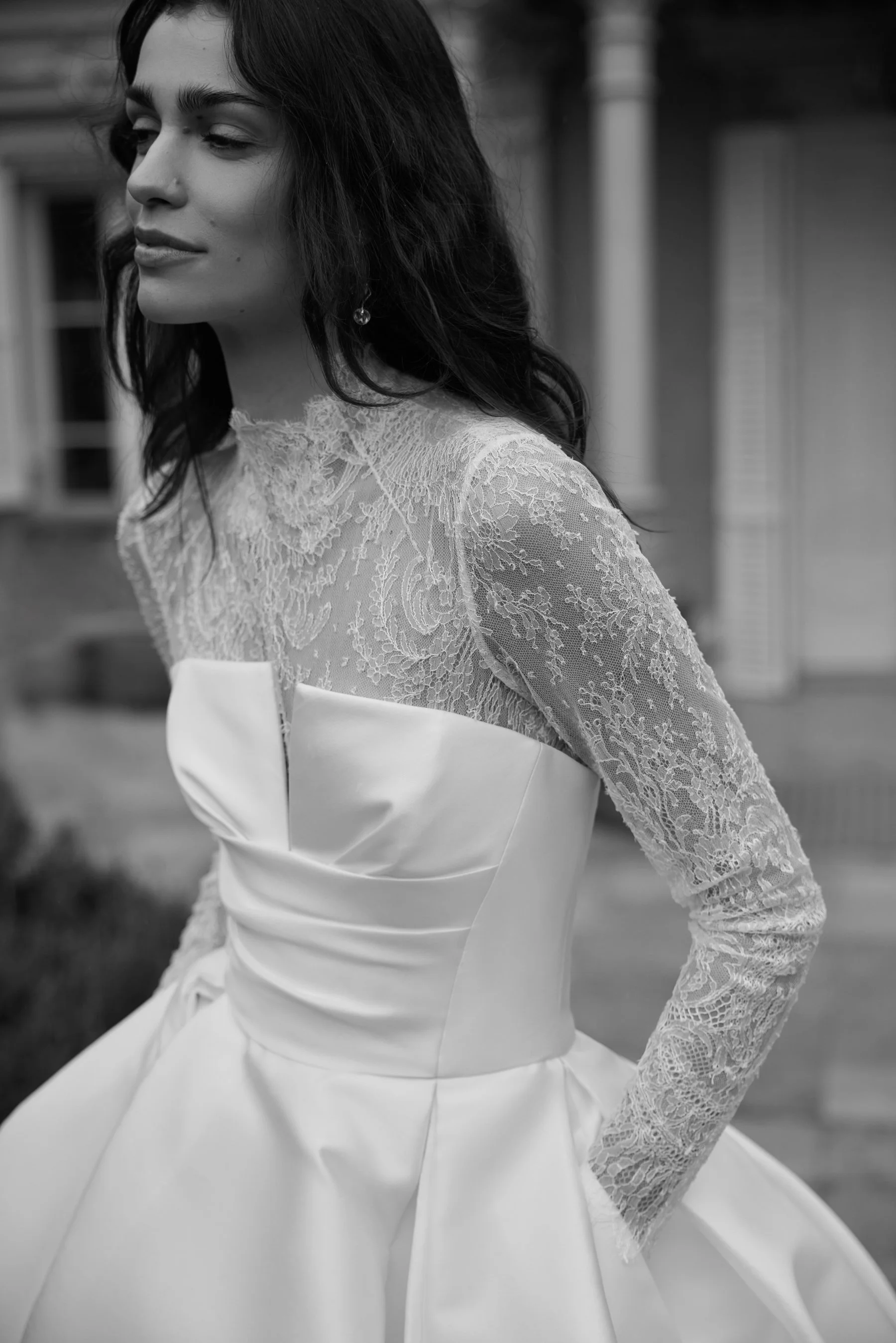 JESUS PEIRO at New York Bridal Market Black and white photo of a woman wearing a wedding dress with lace details on the sleeves and upper chest, looking to the side with her eyes closed, standing outdoors.