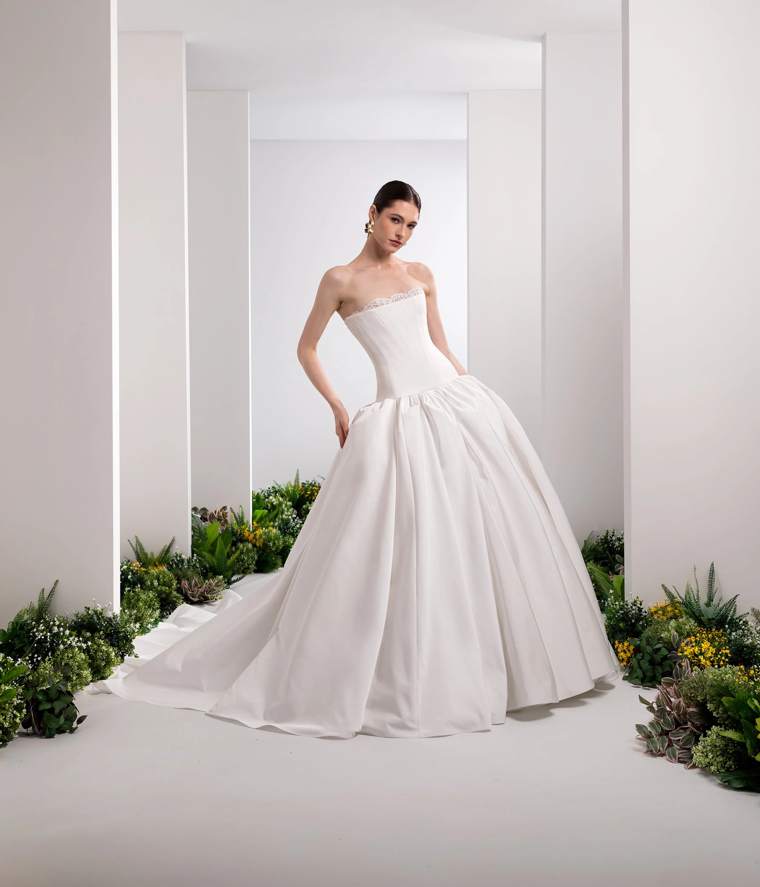 Valentini Spose at New York Bridal Market.  A woman in a white strapless wedding gown posing with her hand on her hip, surrounded by green and yellow plants in a minimalist white setting.